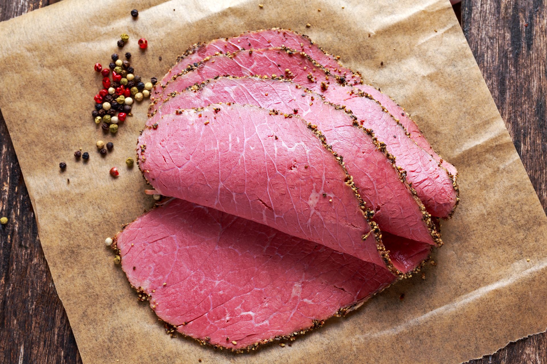 Sliced corned beef on brown paper, sprinkled with peppercorns, on a wooden surface.