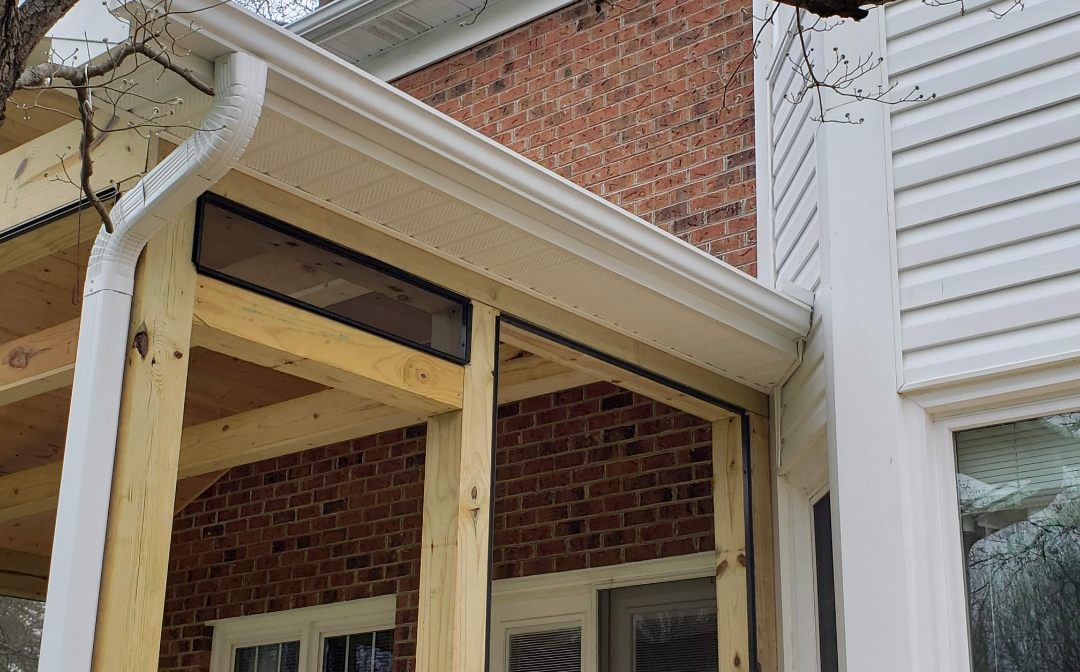 Construction of a wooden porch with white trim, attached to a brick house and a siding-covered side wall.