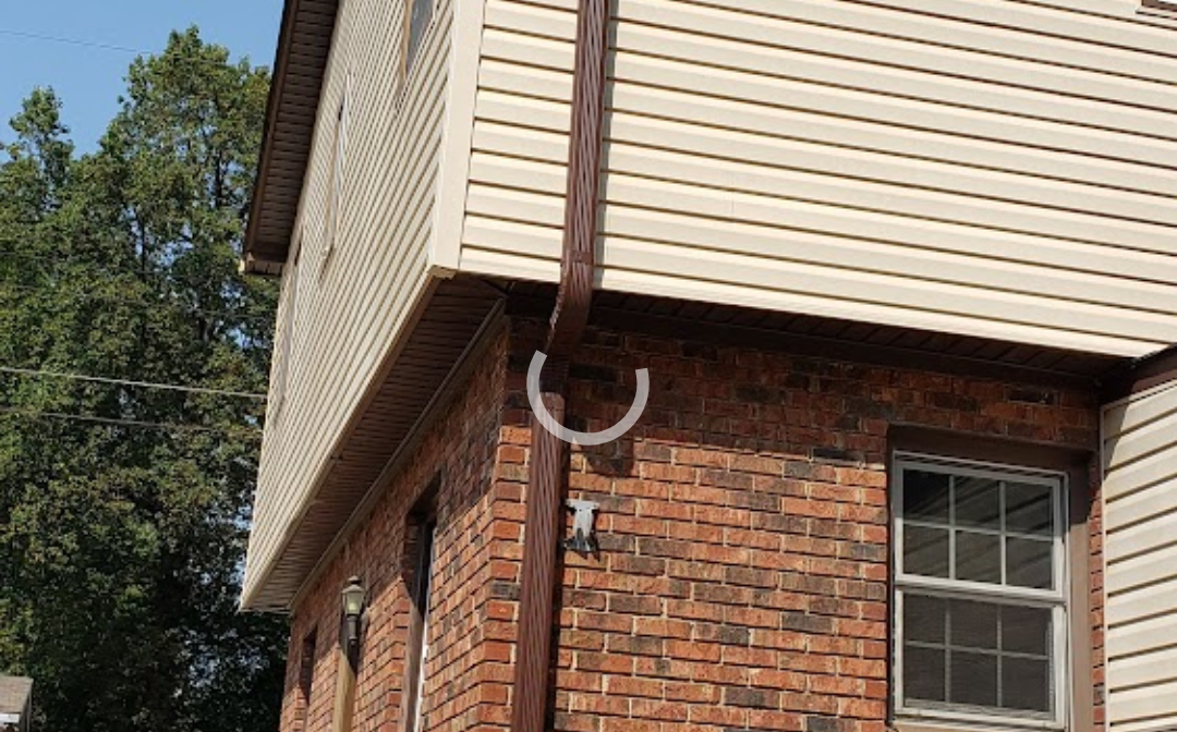 Brown gutter system on a two-story building with tan siding above red brick. A white arch is on the wall.