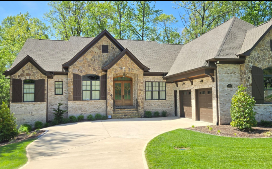 Stone-walled house with brown shutters, garage, and driveway on a sunny day, surrounded by green grass and trees.