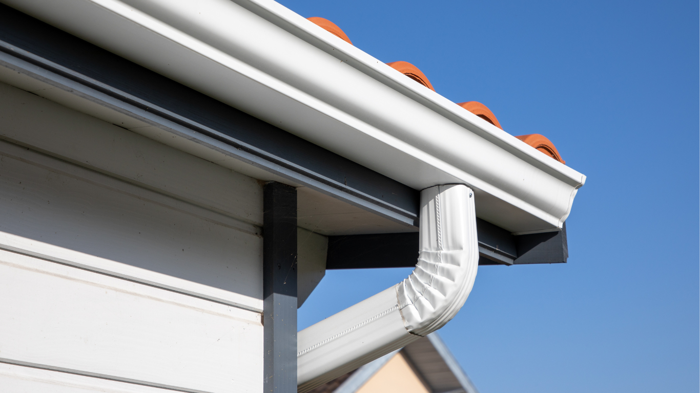 White house gutter with downspout, blue sky, and red tile roof.