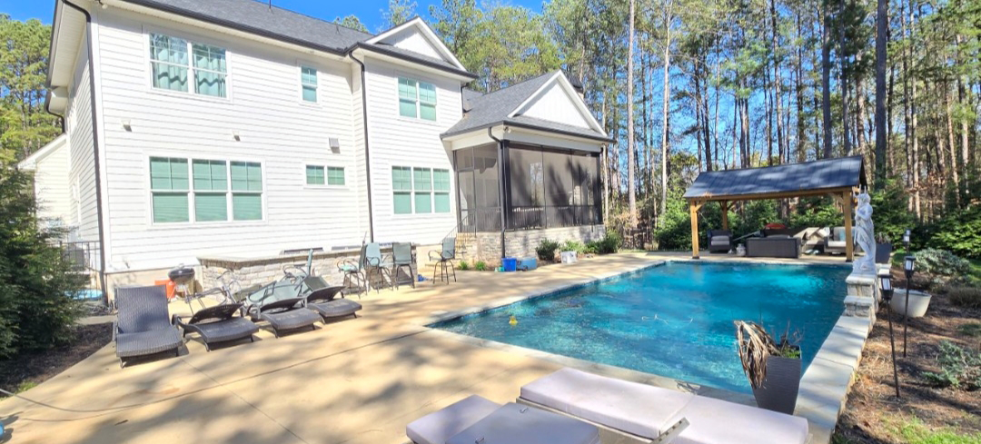 White house with a pool in the backyard, surrounded by trees and a seating area.