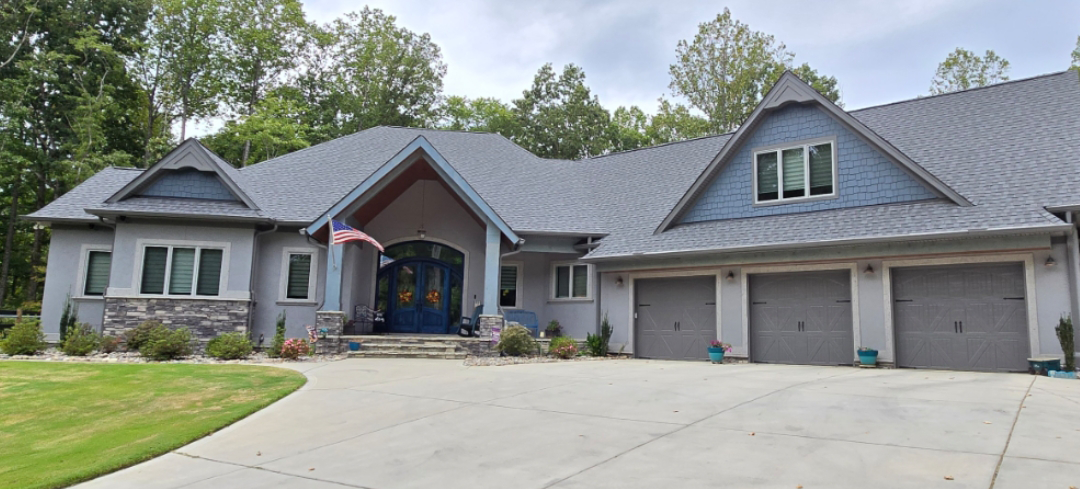Large, modern, grey house with blue accents and three-car garage, set against a backdrop of green trees and a blue sky.