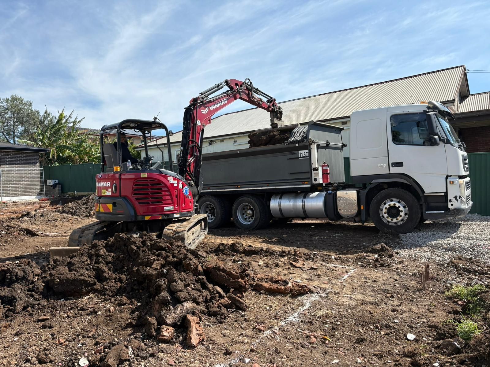 Woman operating a yellow JCB mini excavator, outdoors near a fence.