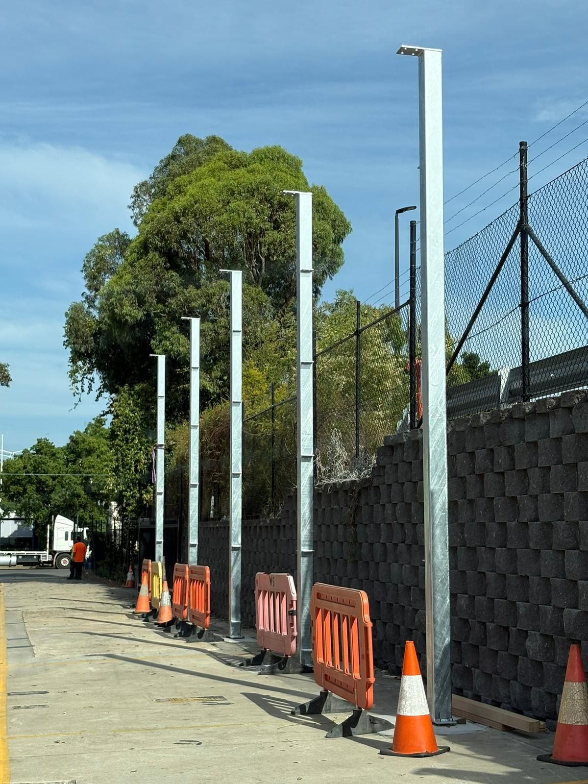 Row of tall metal light poles along a concrete walkway, behind safety barriers and a stone wall.