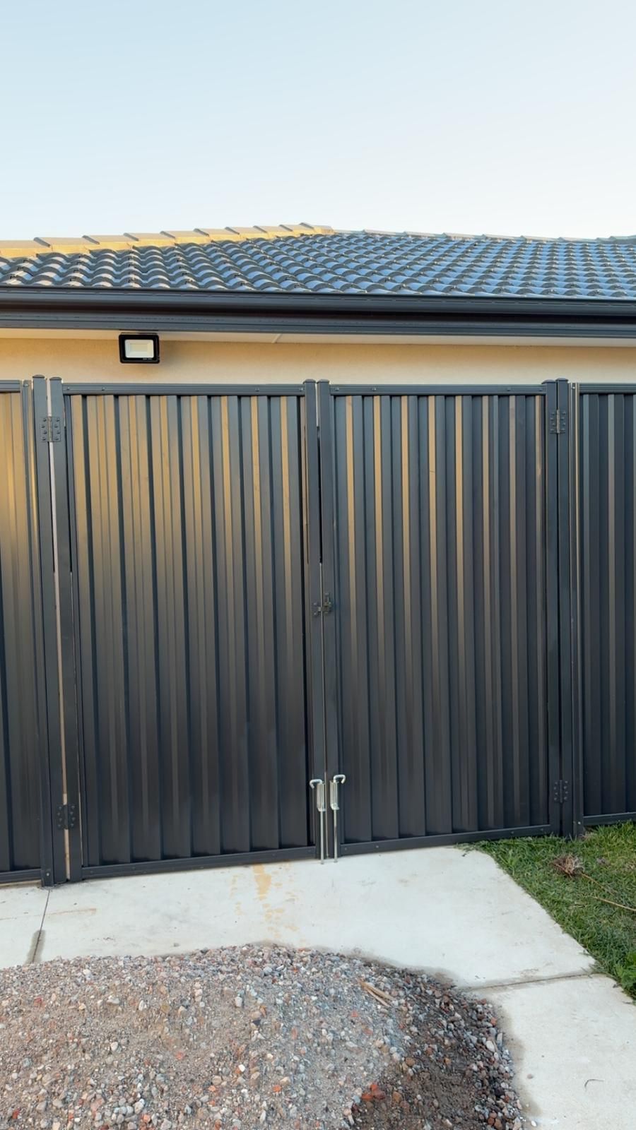 Gray metal gate with vertical ridges in front of a house, set on a concrete and gravel surface.