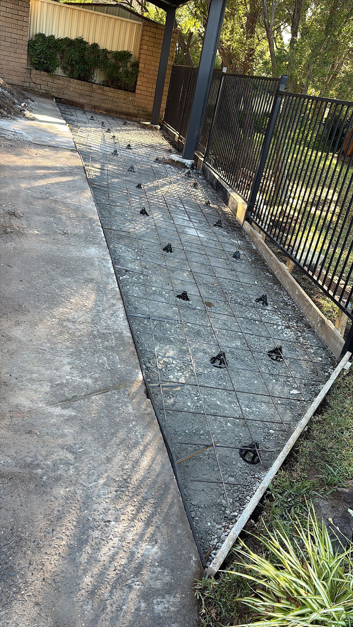 Narrow gravel bed next to concrete walkway and decorative metal fence, with green plants.