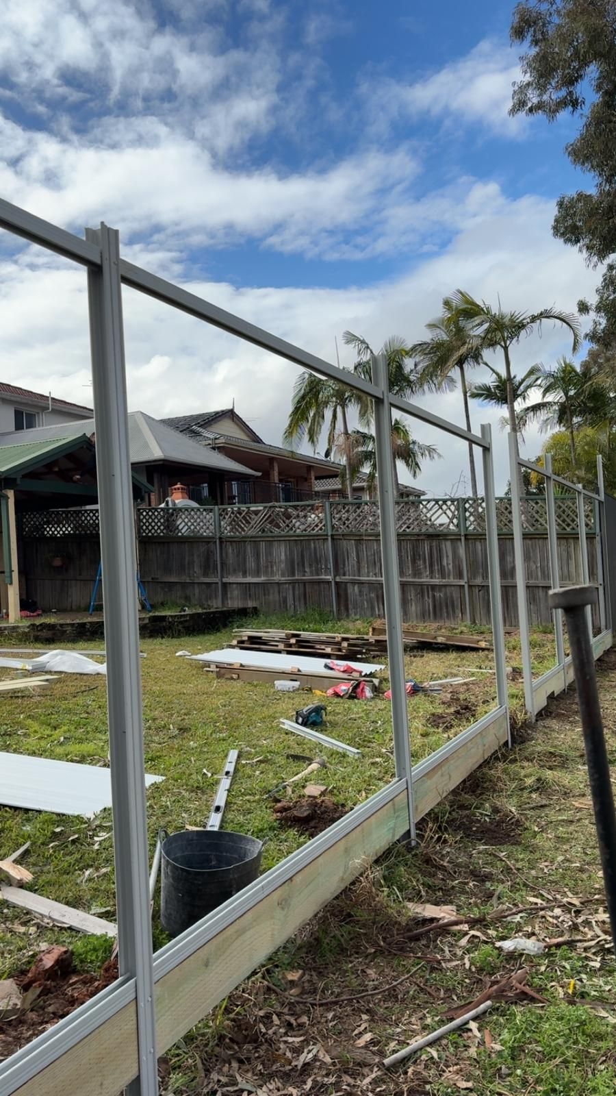 Fence construction: metal frame erected on wooden base, in backyard with house and trees visible.