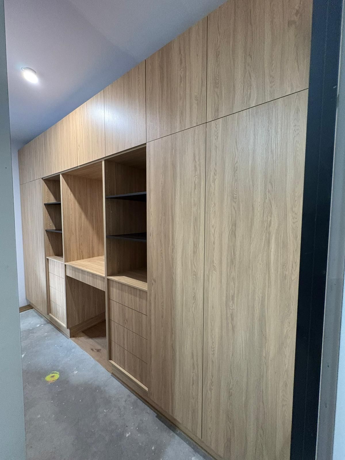 Wooden built-in cabinets with open shelves and a desk area, in a room with unfinished flooring.