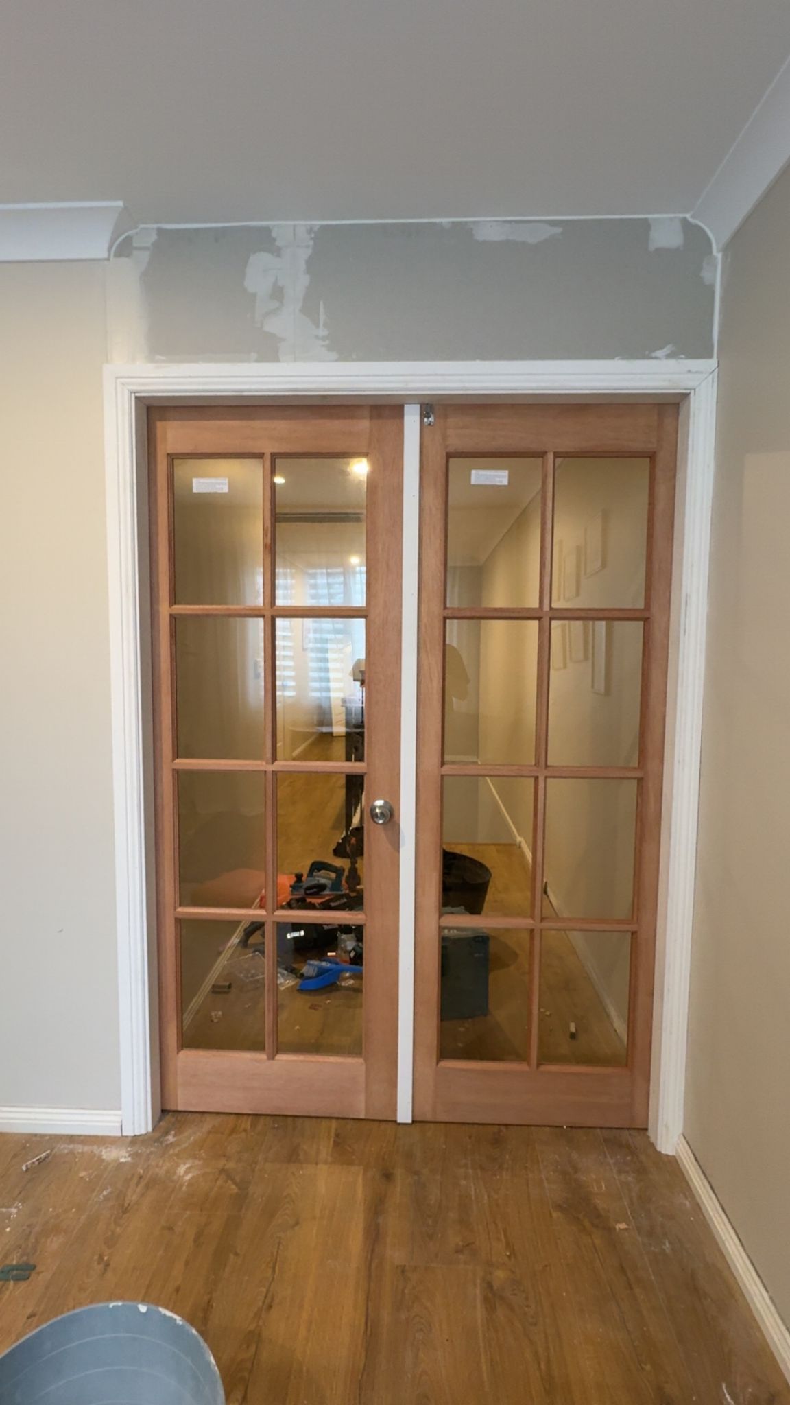 Double wooden doors with glass panes, framed by white trim. Interior setting, tools visible in the hallway.