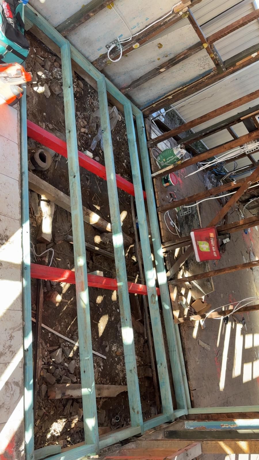 Wooden frame structure under construction with exposed ground and overhead metal roofing.