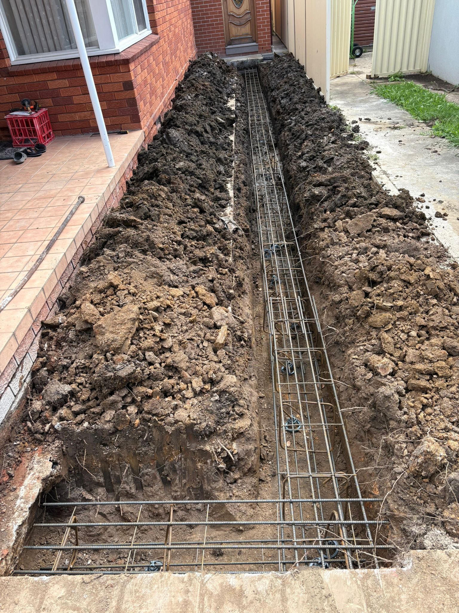 Dug trench with rebar framework, alongside a brick building, ready for concrete.