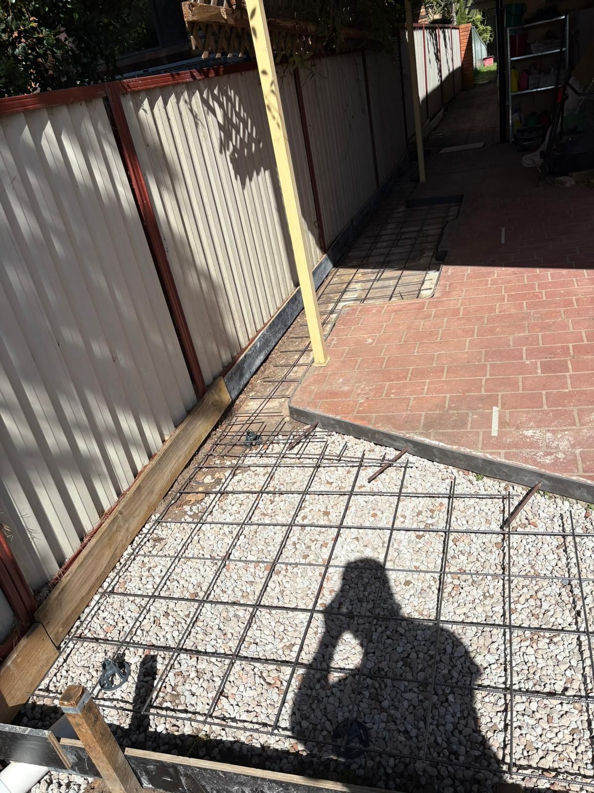 Construction site: a wire grid for concrete sits on gravel. A shadow of a person. Next to a metal fence and brick patio.