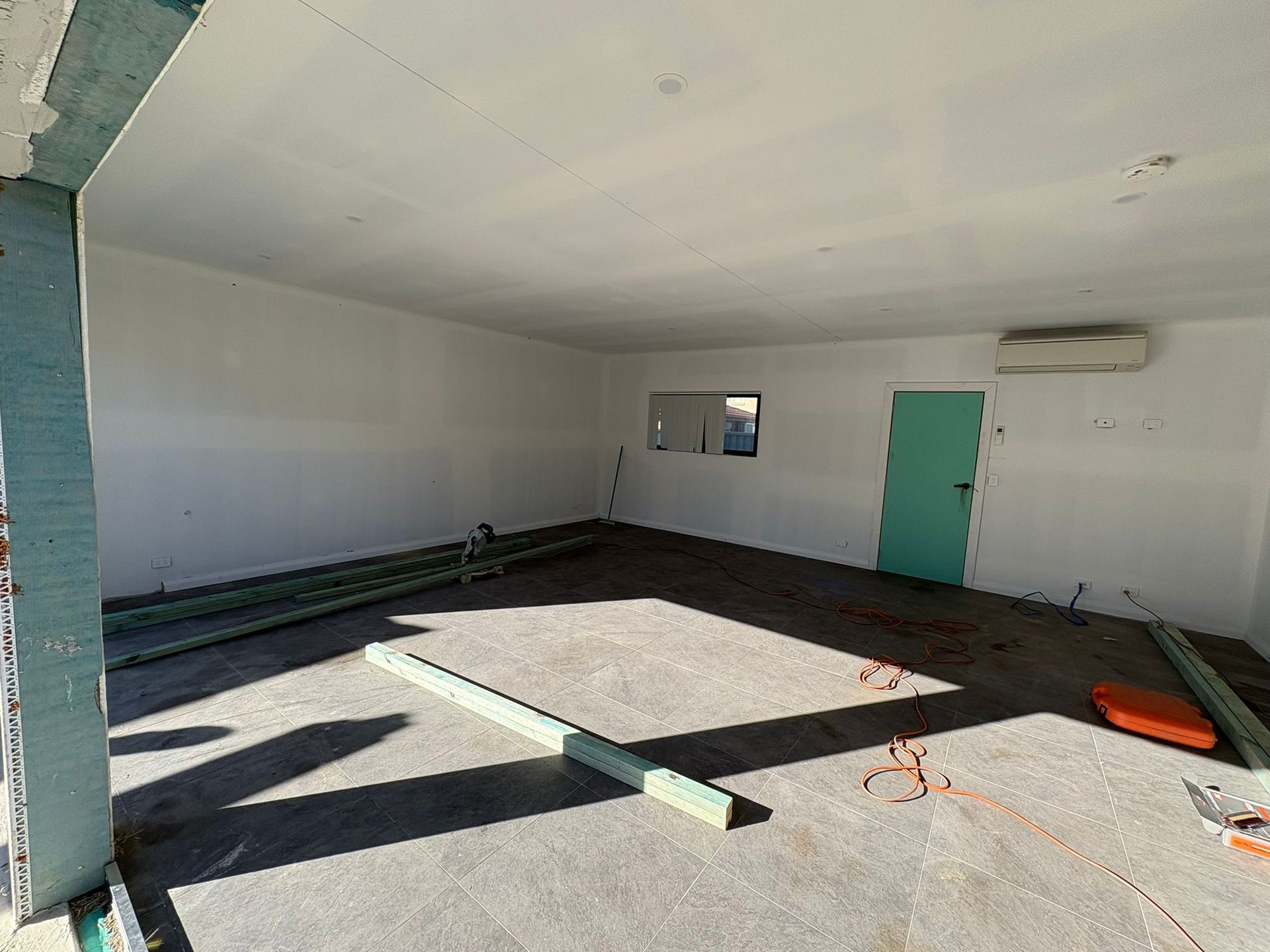 Interior room under construction with white walls, green door, and exposed concrete floor.