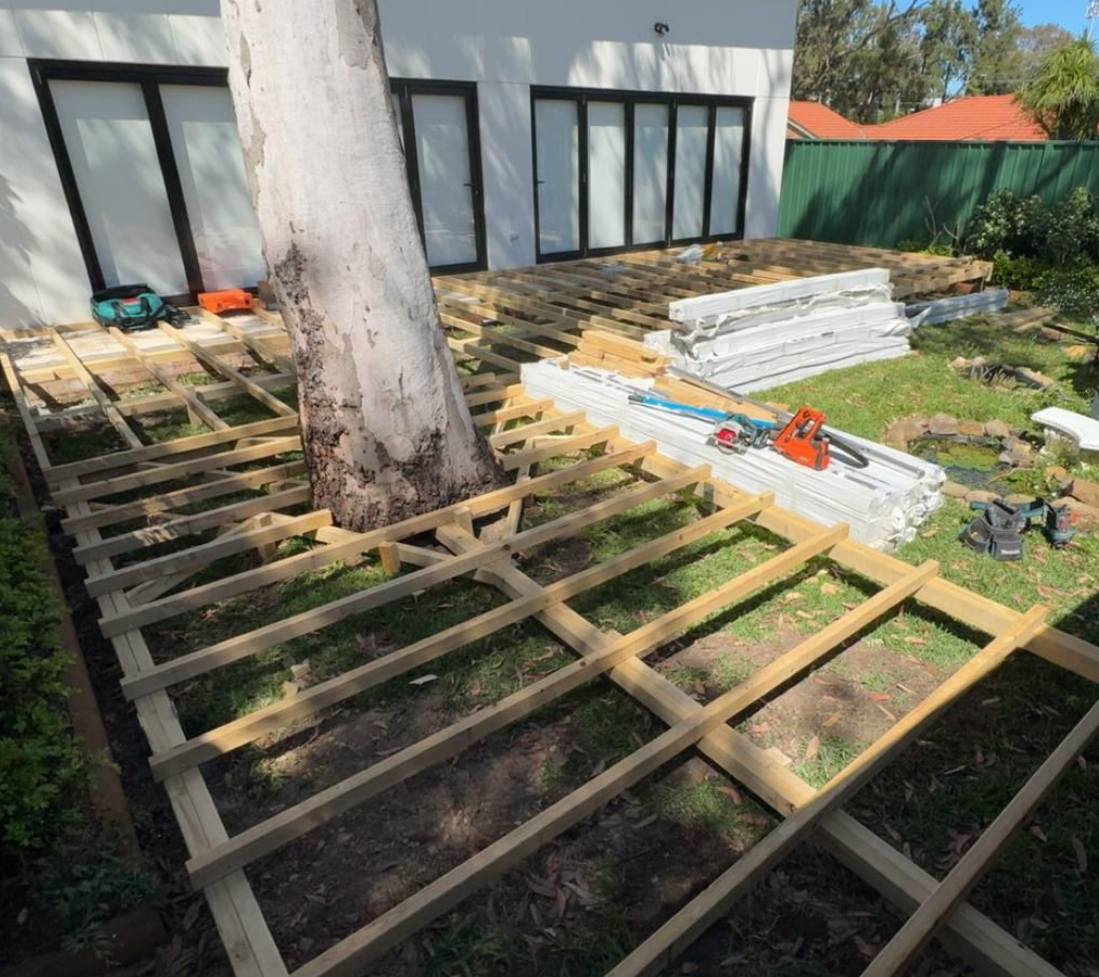 Deck construction in progress around a large tree trunk; lumber frame laid on grass near a white house.