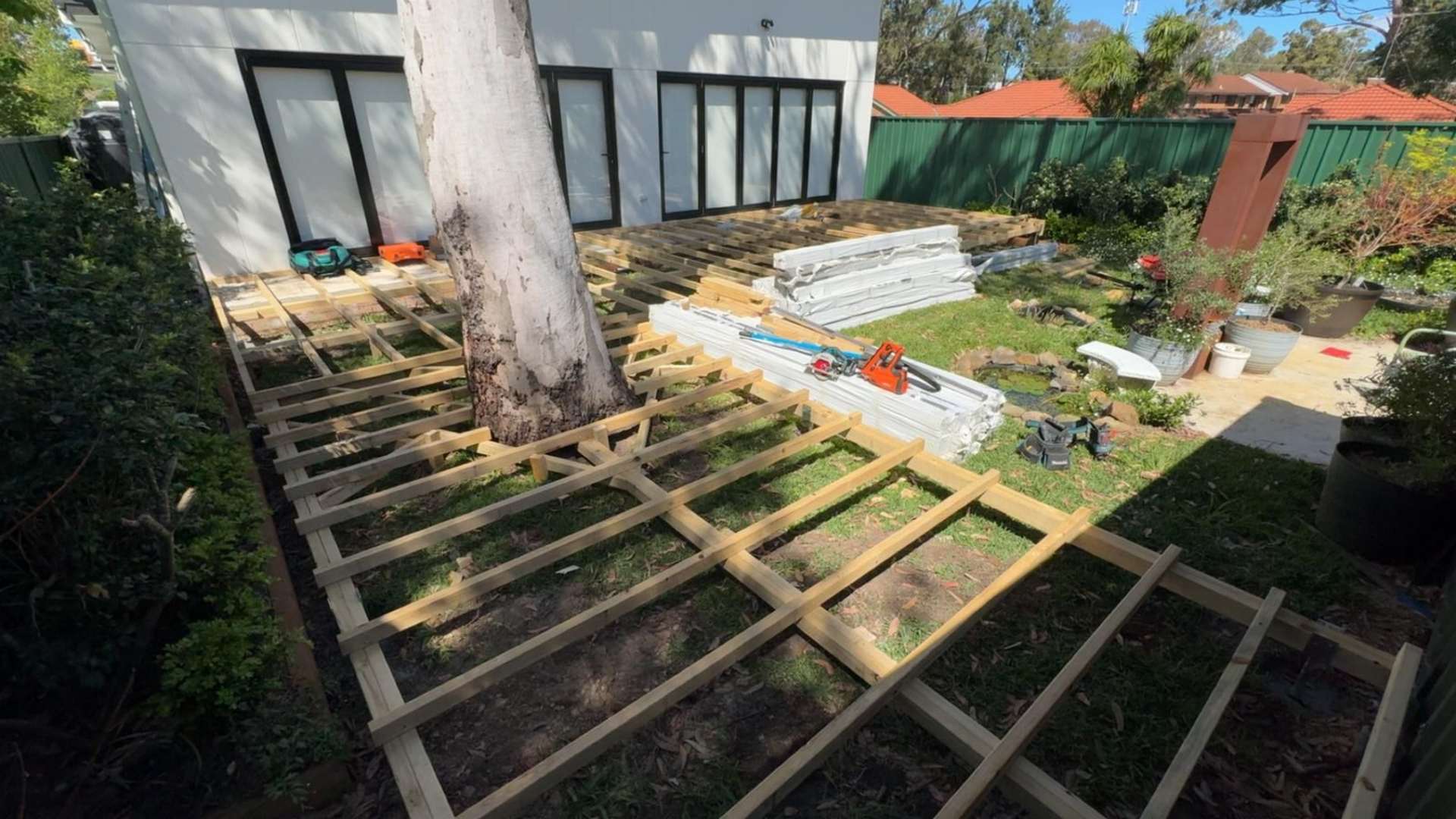 Wooden deck frame under construction around a large tree in a backyard.