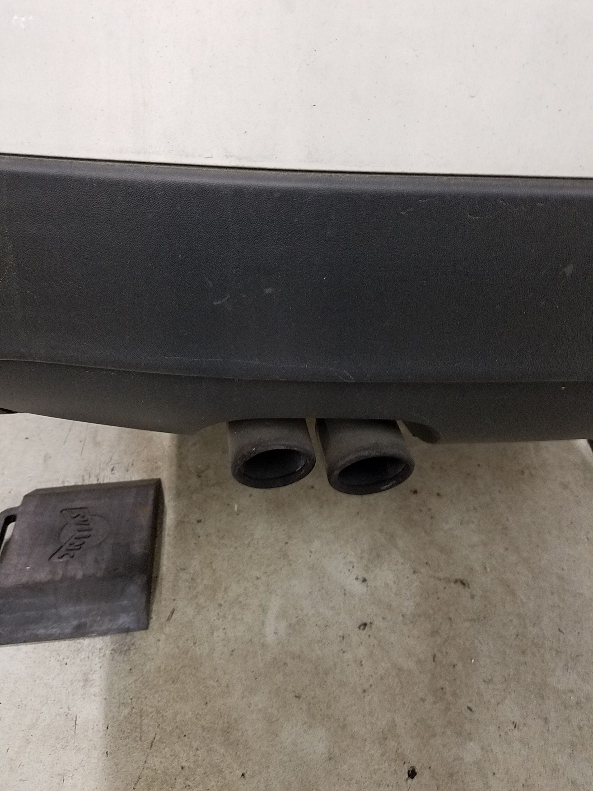 Black car bumper with dual exhaust pipes. A black, square object sits on the floor. | Autobahn Performance Inc