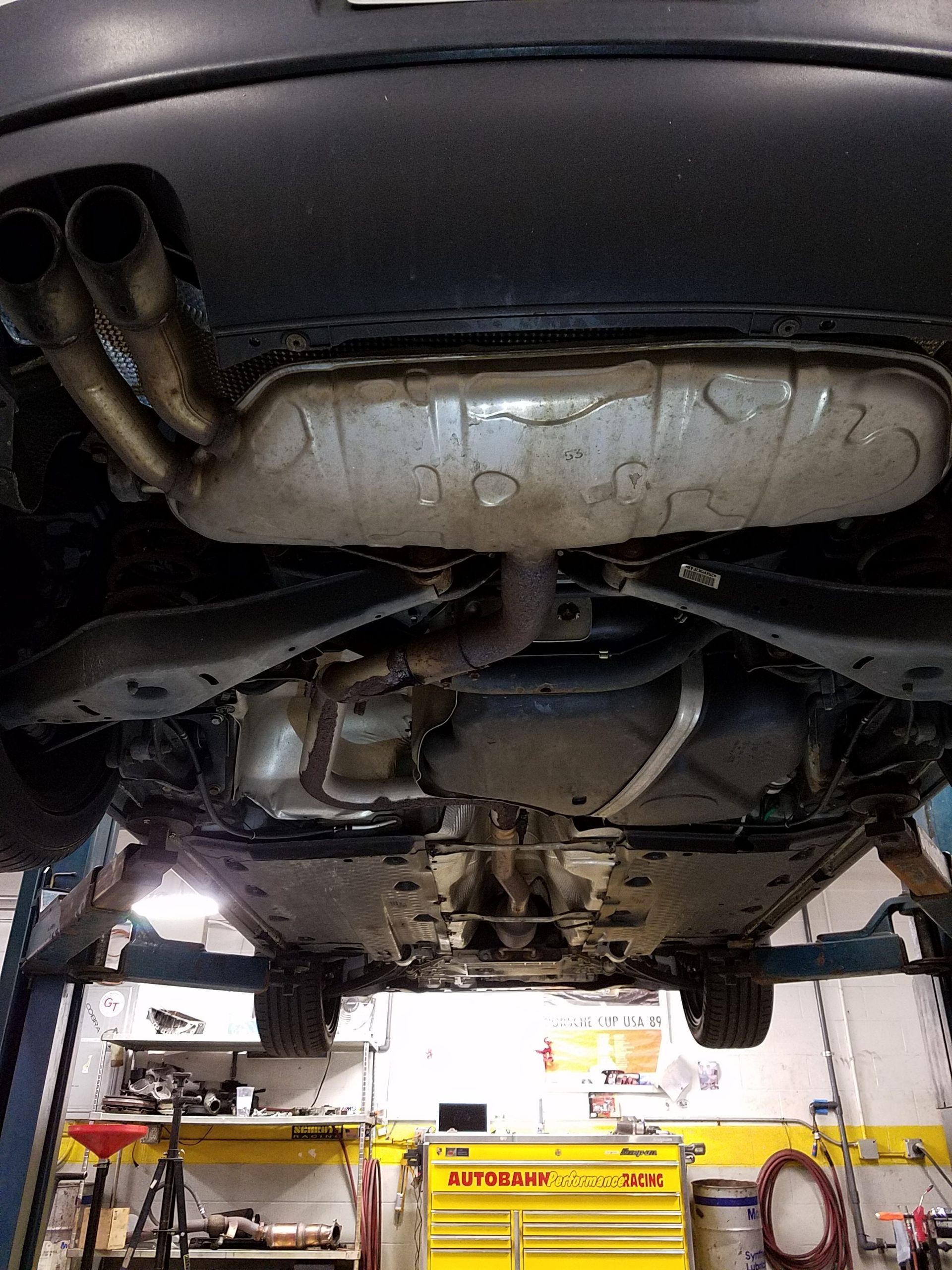 Underside of a car on a lift; exhaust system visible, including muffler and dual tailpipes. Setting is a garage. | Autobahn Performance Inc