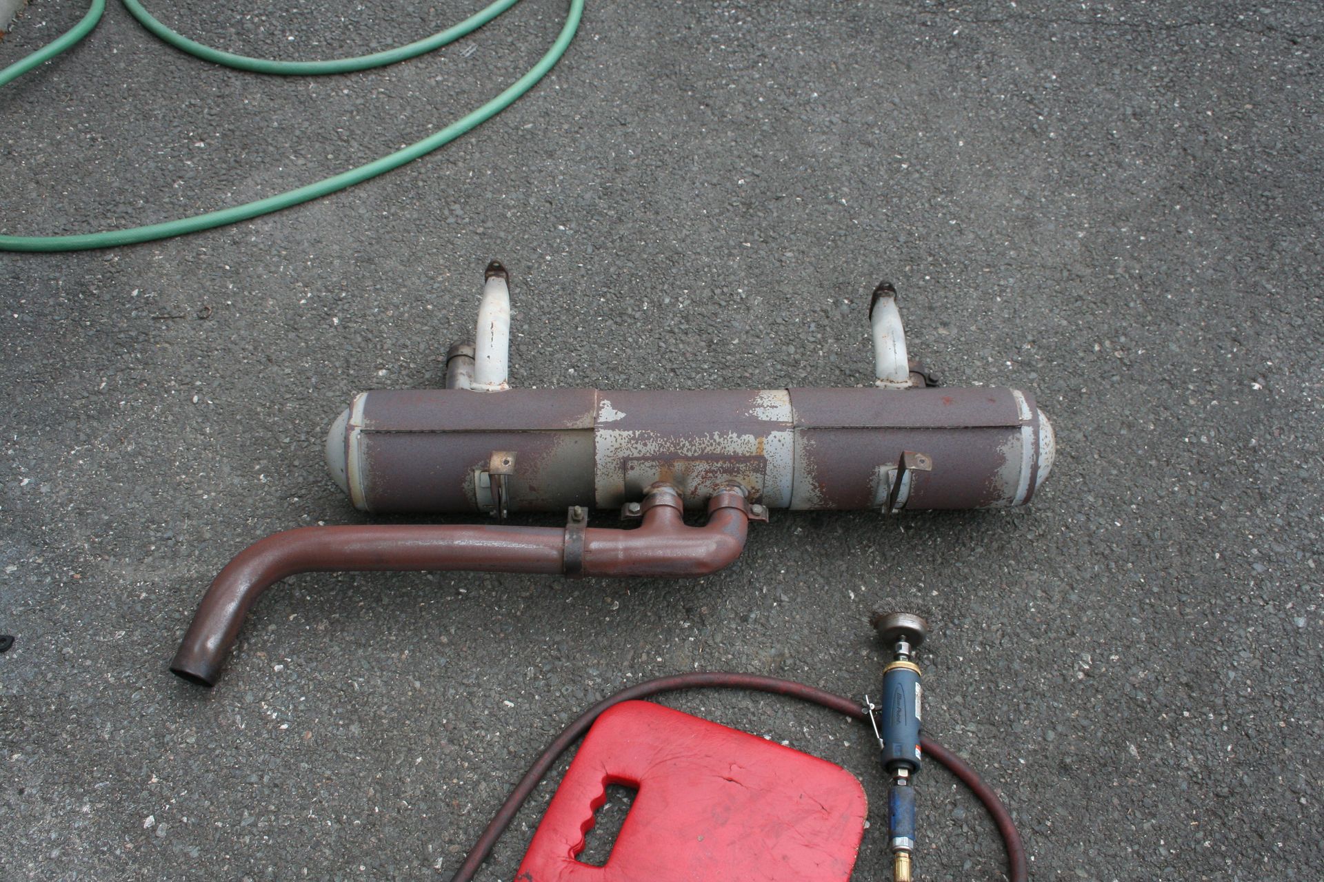 Brown and gray muffler with two vertical tubes, on gray concrete, with a red container and hose. | Autobahn Performance Inc