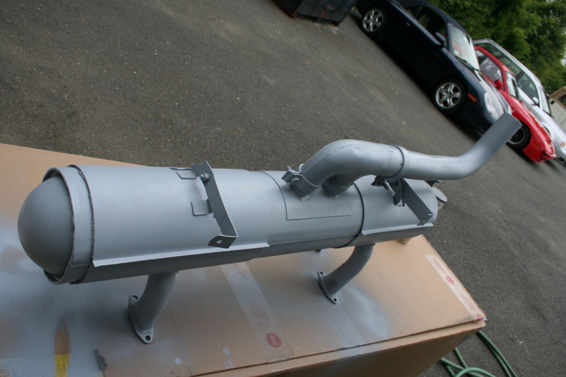 Gray painted car exhaust on a cardboard box, with a curved pipe and support brackets, outdoor setting with cars. | Autobahn Performance Inc