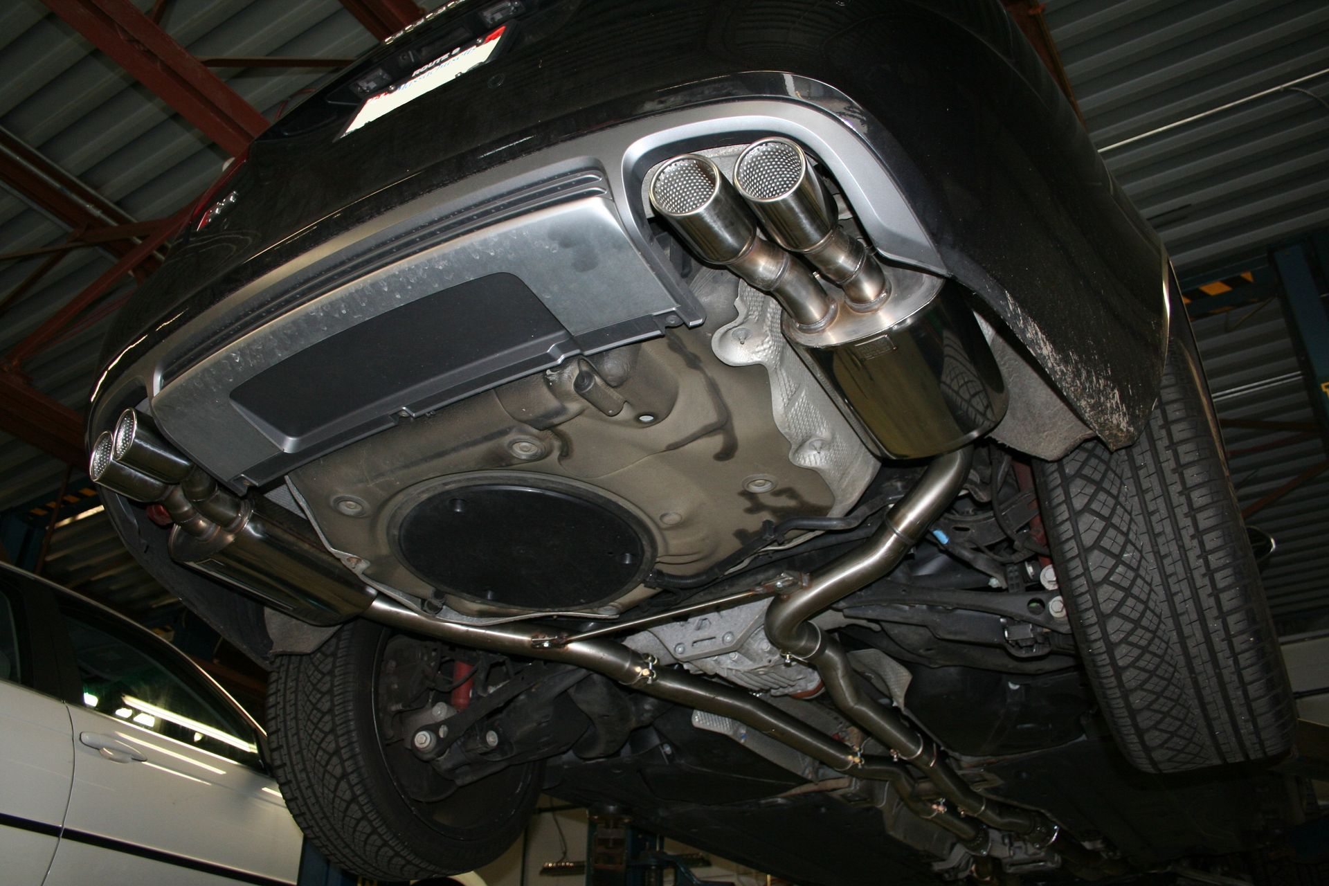 Undercarriage of a black car, showing exhaust system and four chrome exhaust tips. Inside a repair shop. | Autobahn Performance Inc