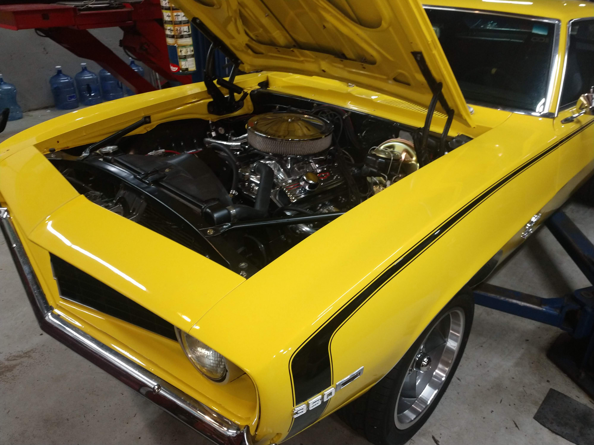 Mechanic working on a car engine in a garage with another car on a lift. | Autobahn Performance Inc