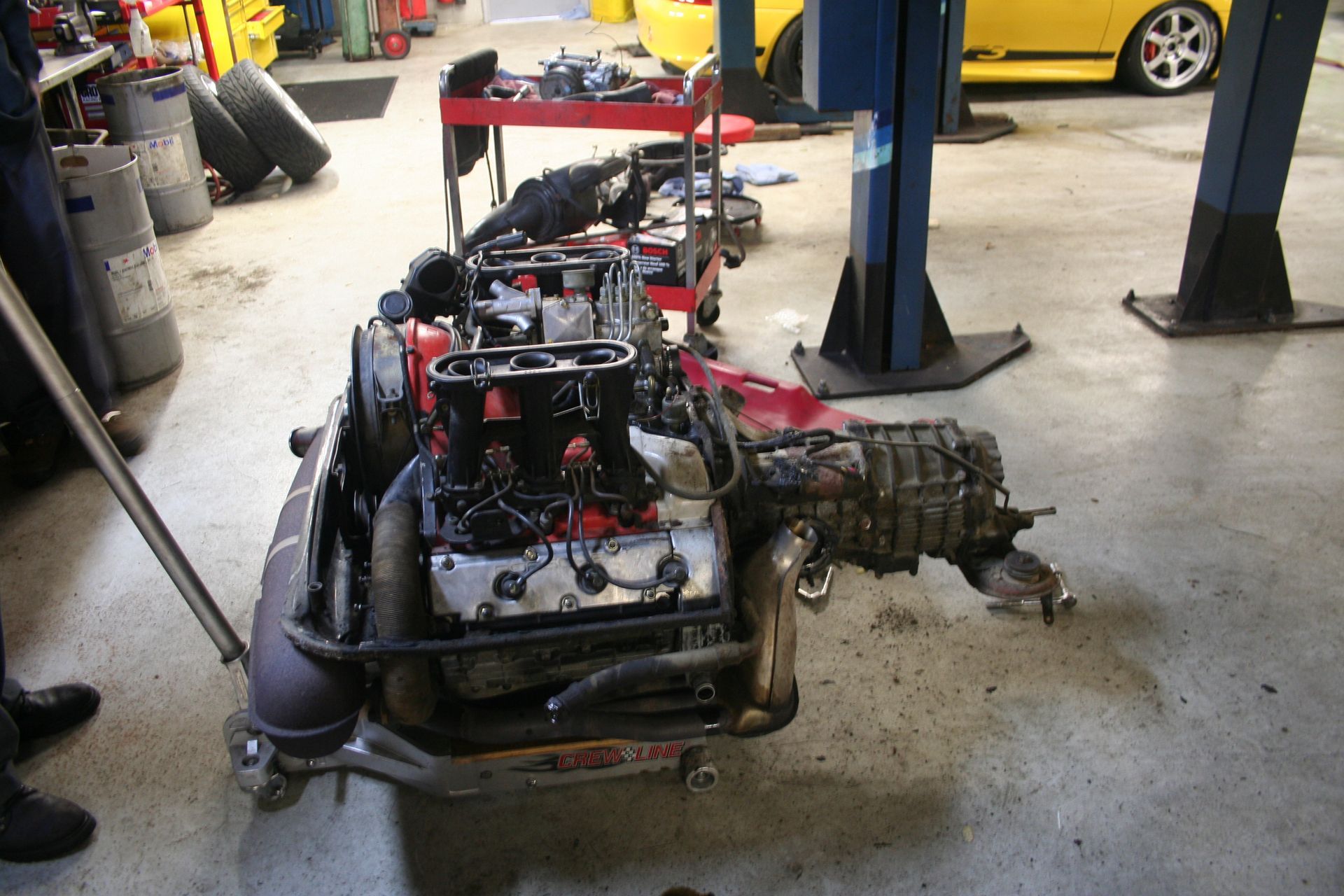 Engine and transmission assembly on a rolling cart in a mechanic's garage with a yellow car in the background. | Autobahn Performance Inc