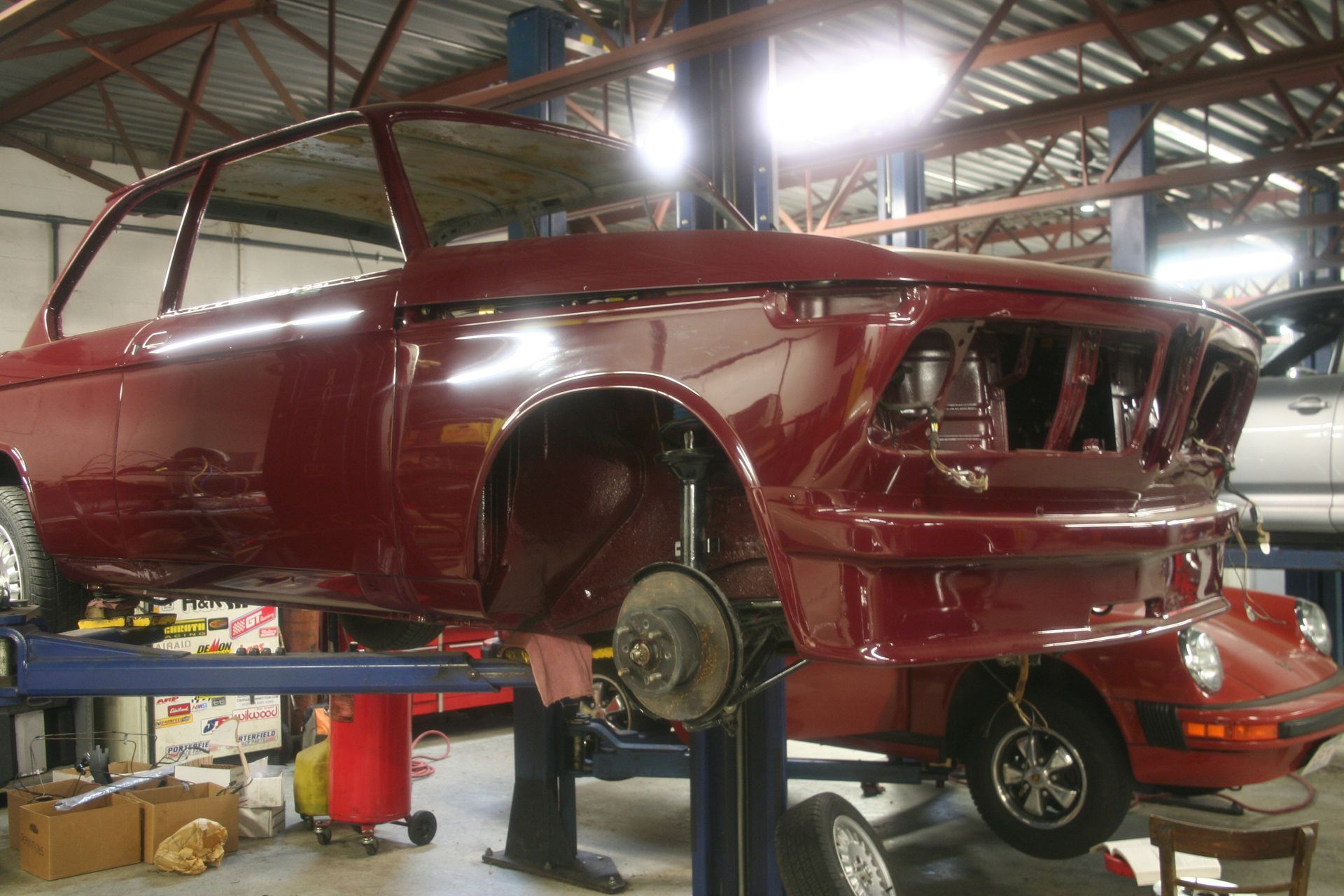 Maroon car on a lift, bodywork in progress. Red brake calipers visible, tires mounted. Workshop setting. | Autobahn Performance Inc