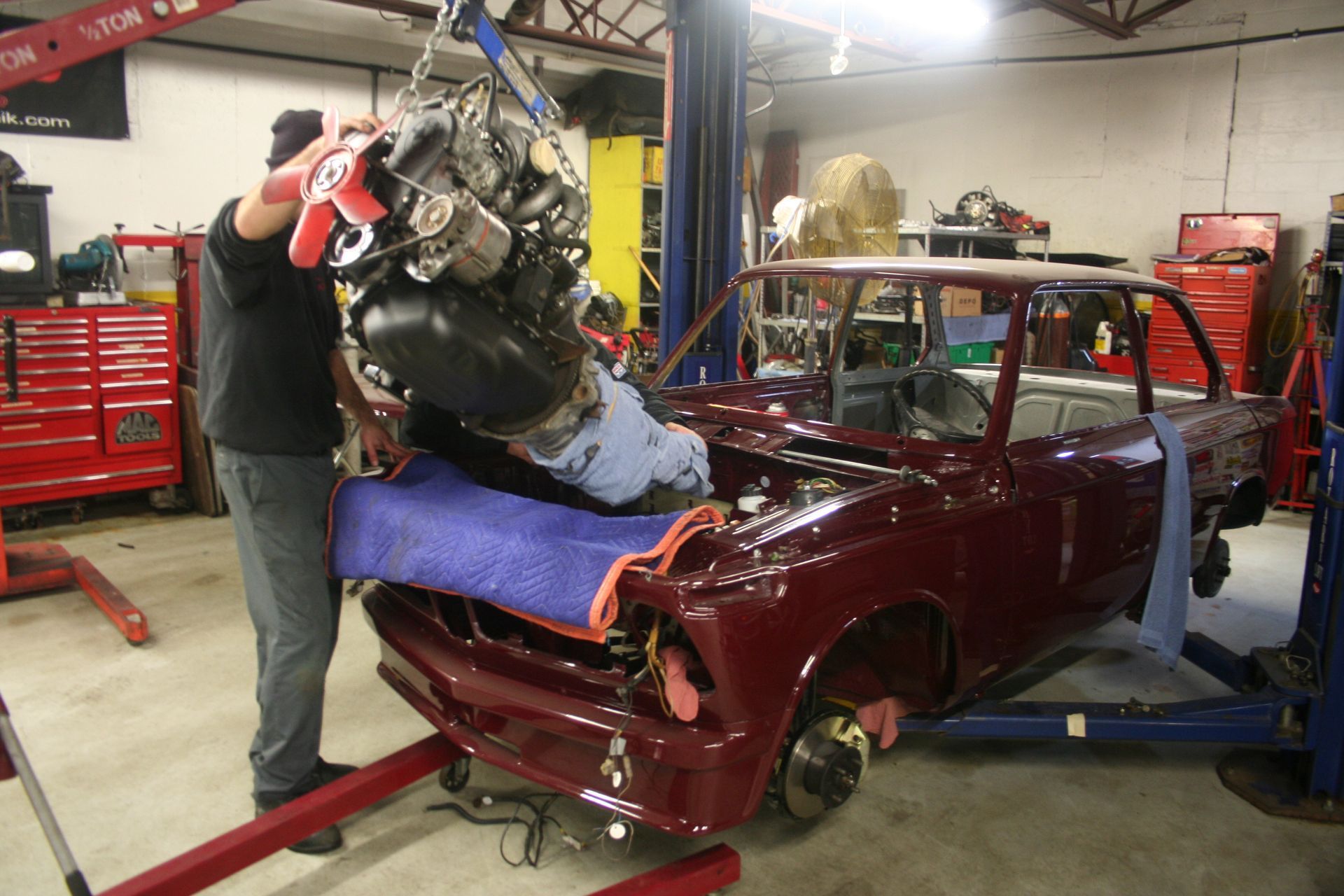 Man installing an engine into a maroon car in a garage with a lift. | Autobahn Performance Inc