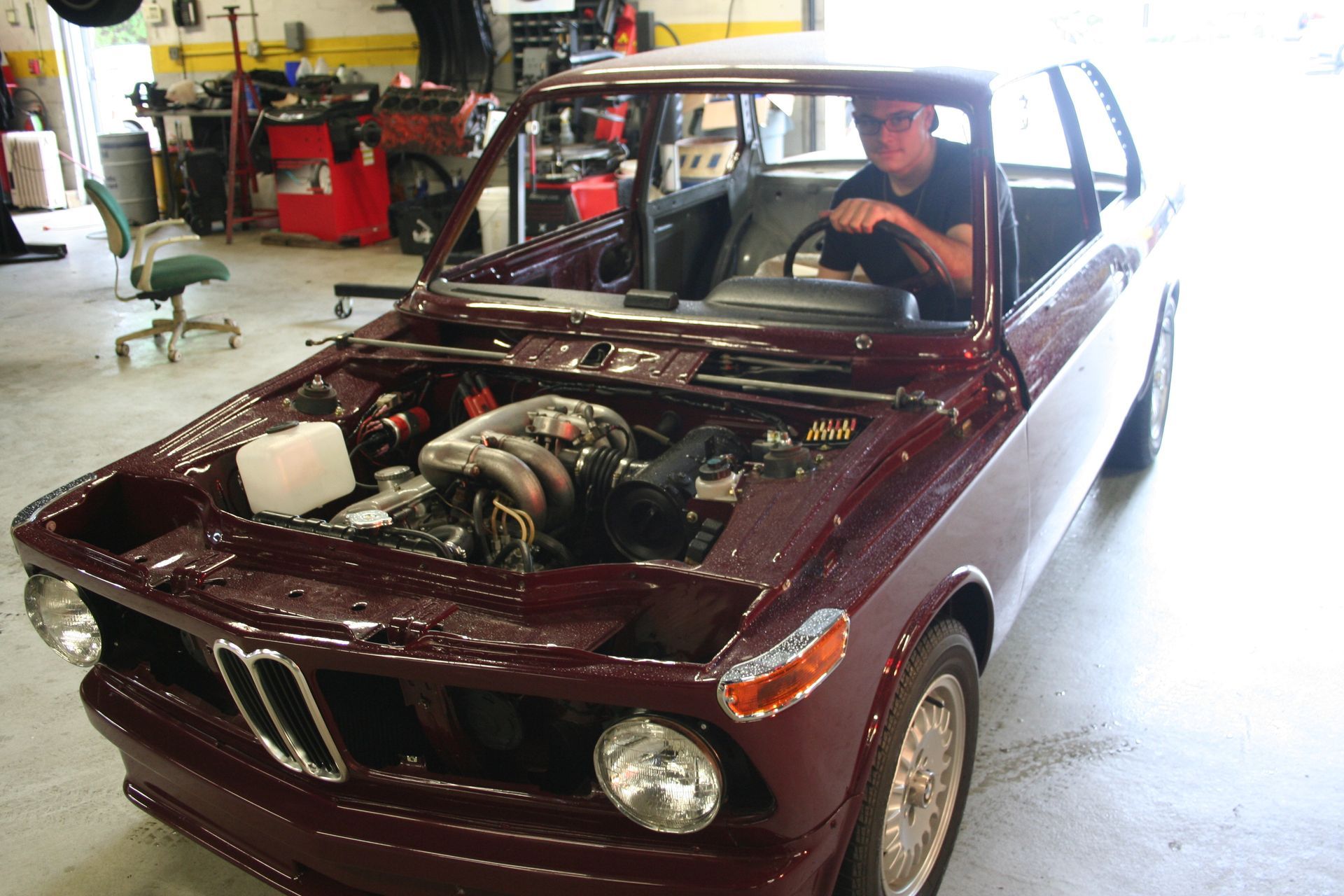 A person sits in a maroon BMW with the hood open, revealing the engine, inside a garage. | Autobahn Performance Inc
