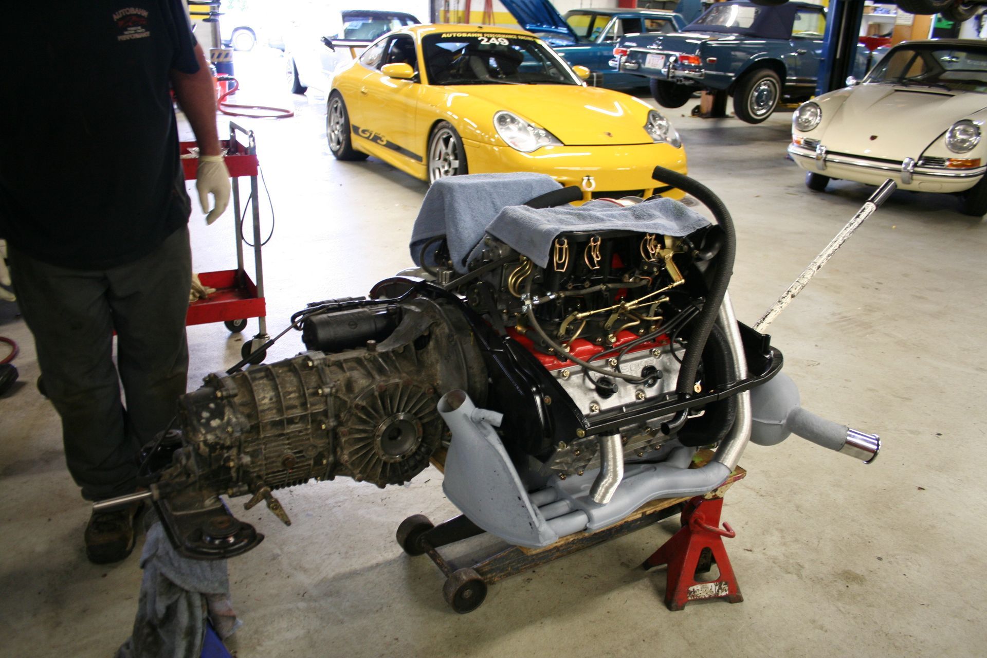 Person holding a car engine and transmission in a repair shop, with yellow and cream Porsche cars in the background. | Autobahn Performance Inc