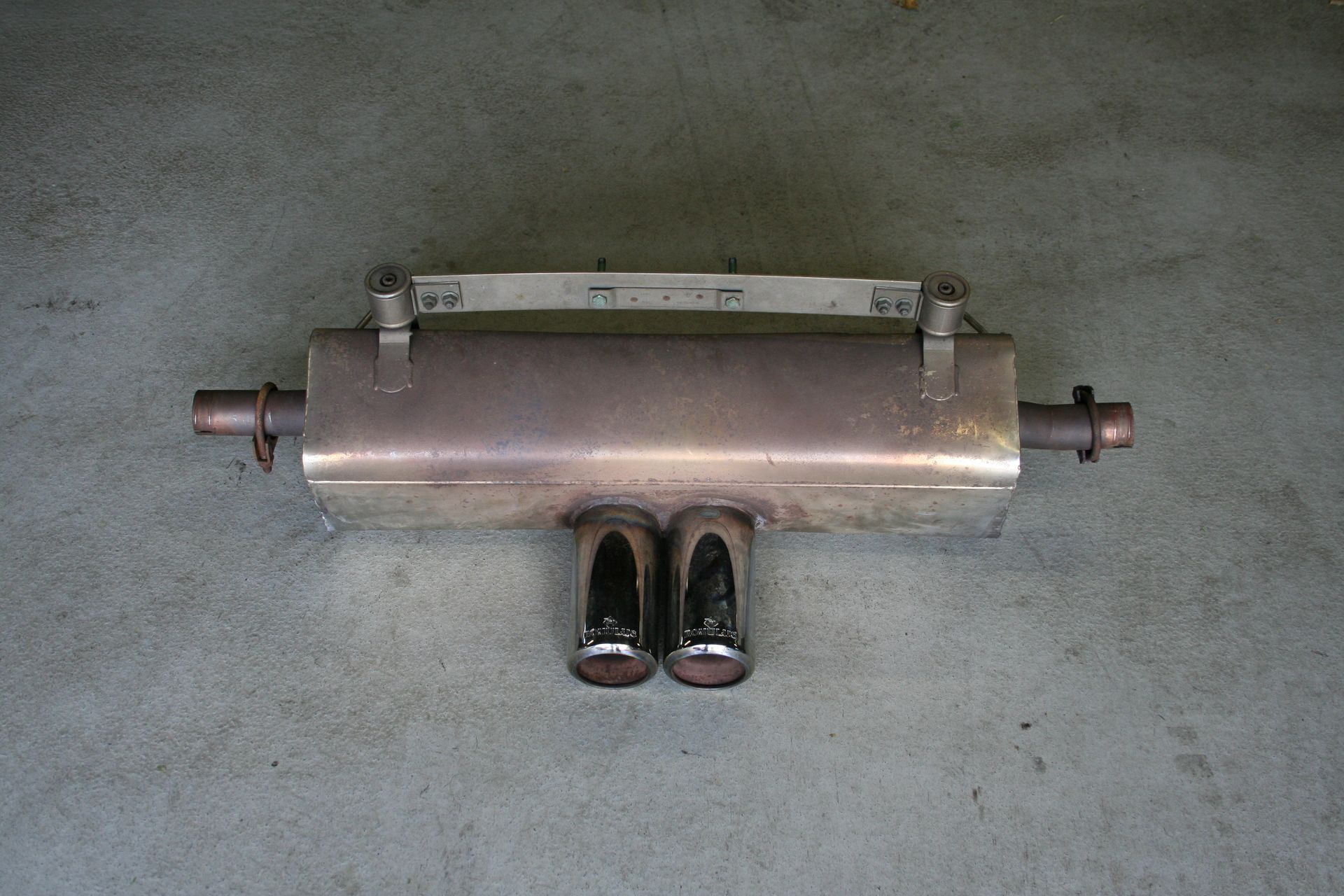 Metal car muffler with two exhaust pipes, mounted on a gray surface. | Autobahn Performance Inc