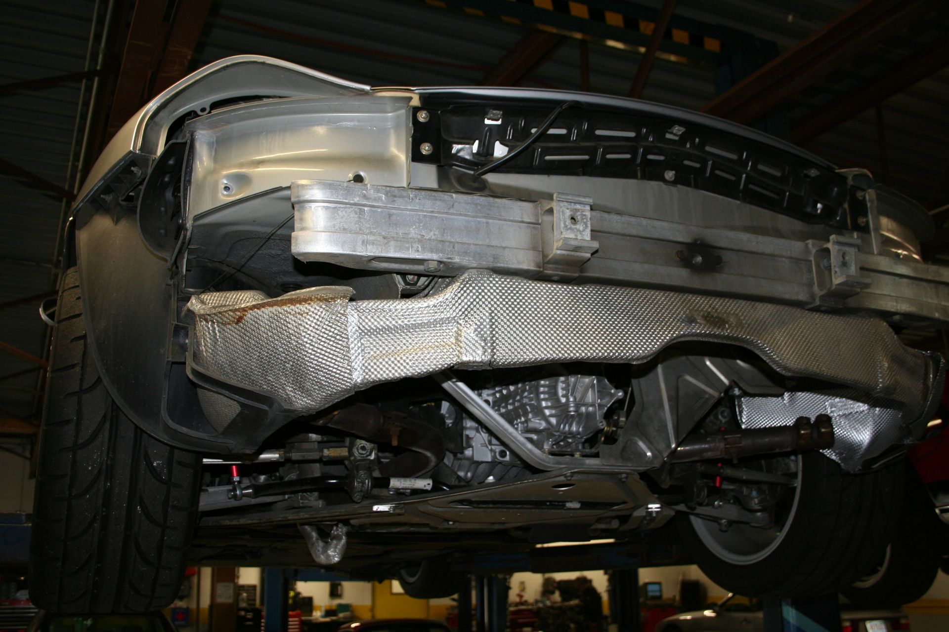Undercarriage of a silver car on a lift, showing suspension, exhaust system, and metal framework. | Autobahn Performance Inc