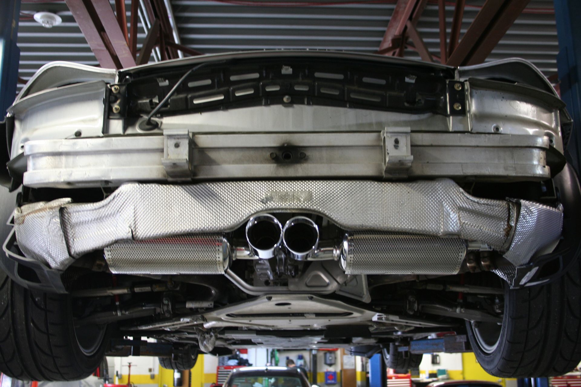 Underside of a car on a lift; silver exhaust pipes, silver heat shields. Black tires visible. Automotive shop setting. | Autobahn Performance Inc