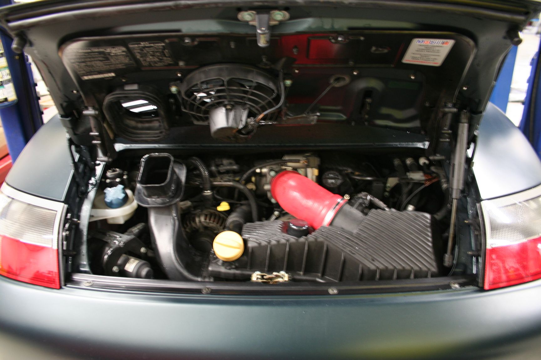 Engine compartment of a teal Porsche, hood open. Features include fan, black air filter, red pipe, and various engine components. | Autobahn Performance Inc