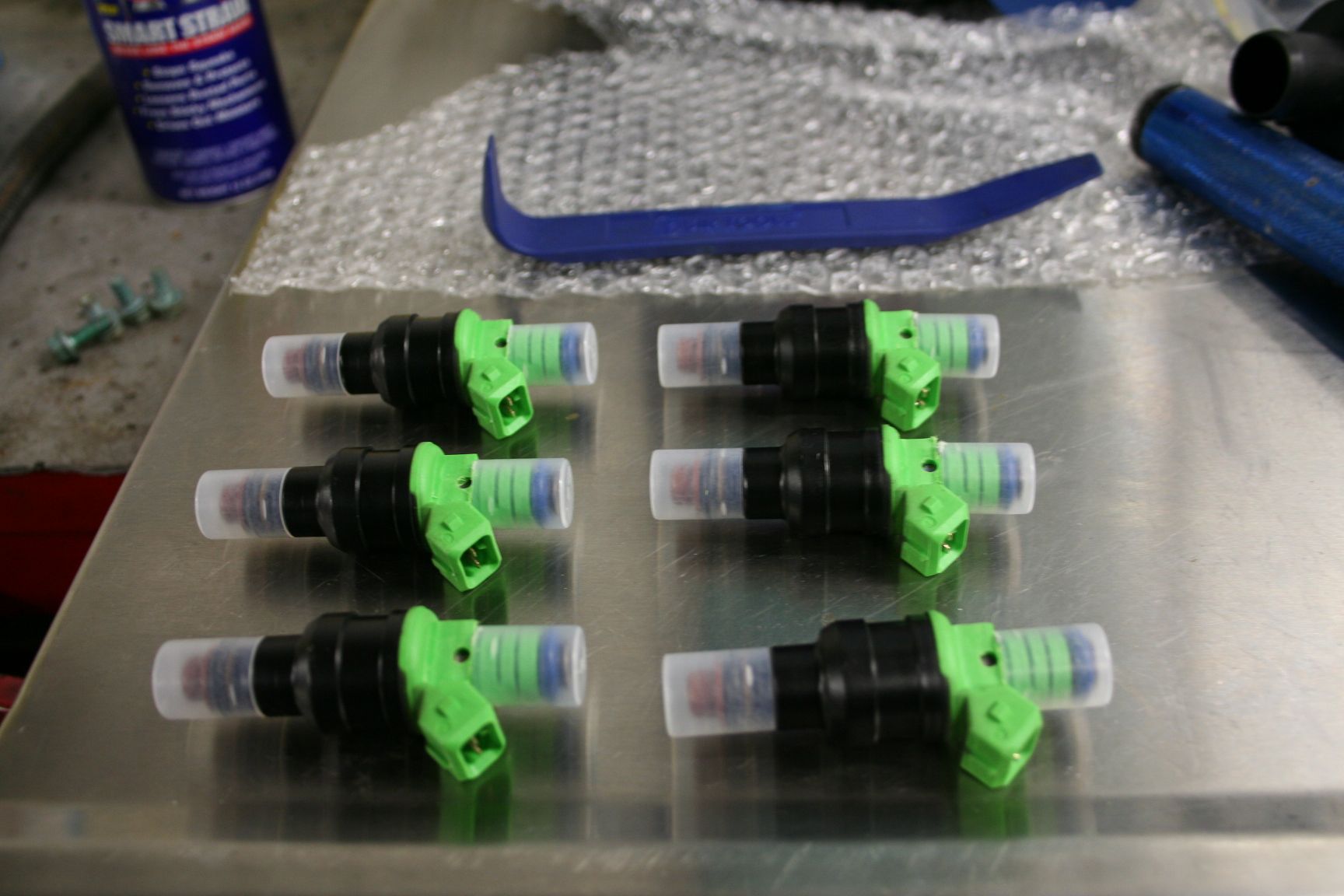 Six fuel injectors with green connectors, on a metal surface. | Autobahn Performance Inc
