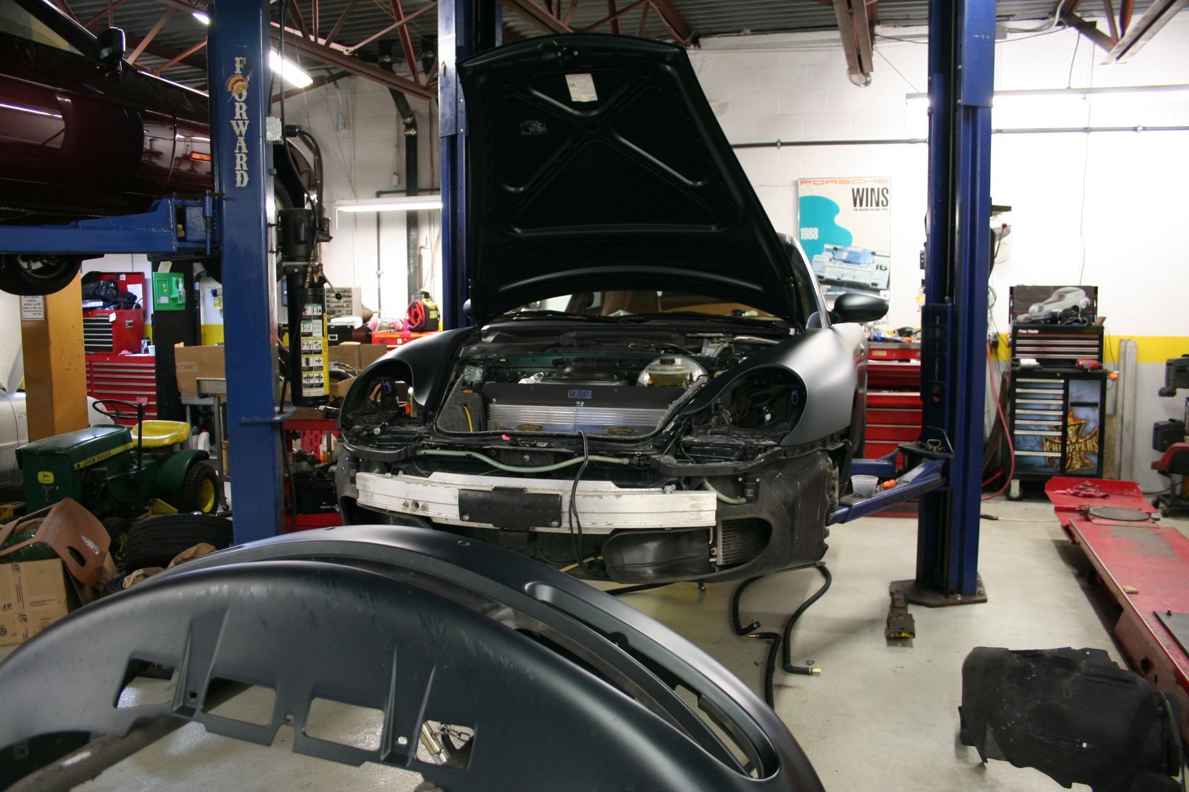 A dark green sports car in a repair shop with the hood open and front bumper removed, a second bumper in the foreground. | Autobahn Performance Inc