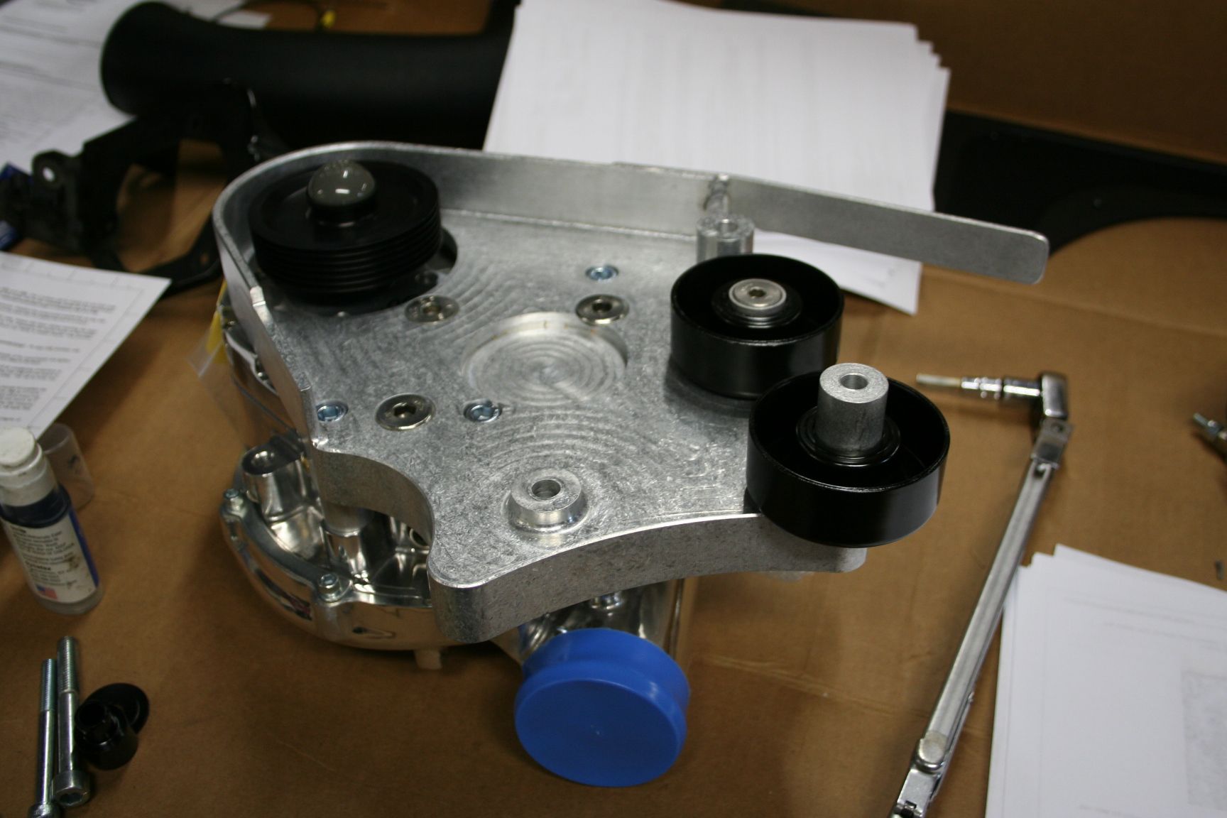 Silver automotive component with black pulleys and blue cap, lying on a table with a tool. | Autobahn Performance Inc