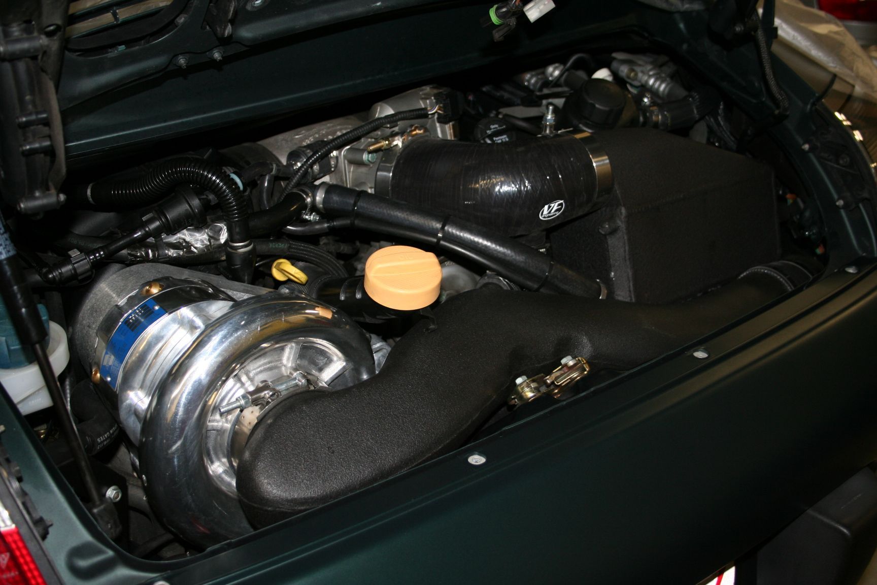 Engine bay of a dark green vehicle with a large turbocharger, black intake, and various components visible. | Autobahn Performance Inc