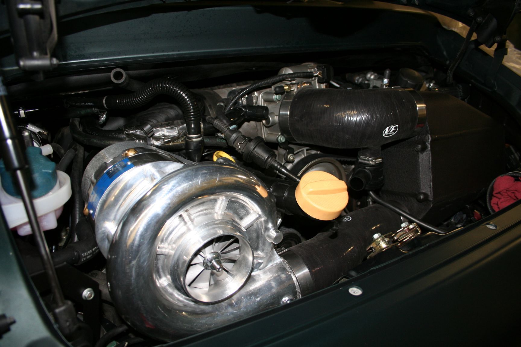 Close-up of a car engine bay, showcasing a large silver supercharger with blue accents. Black tubing and a yellow cap are also visible. | Autobahn Performance Inc