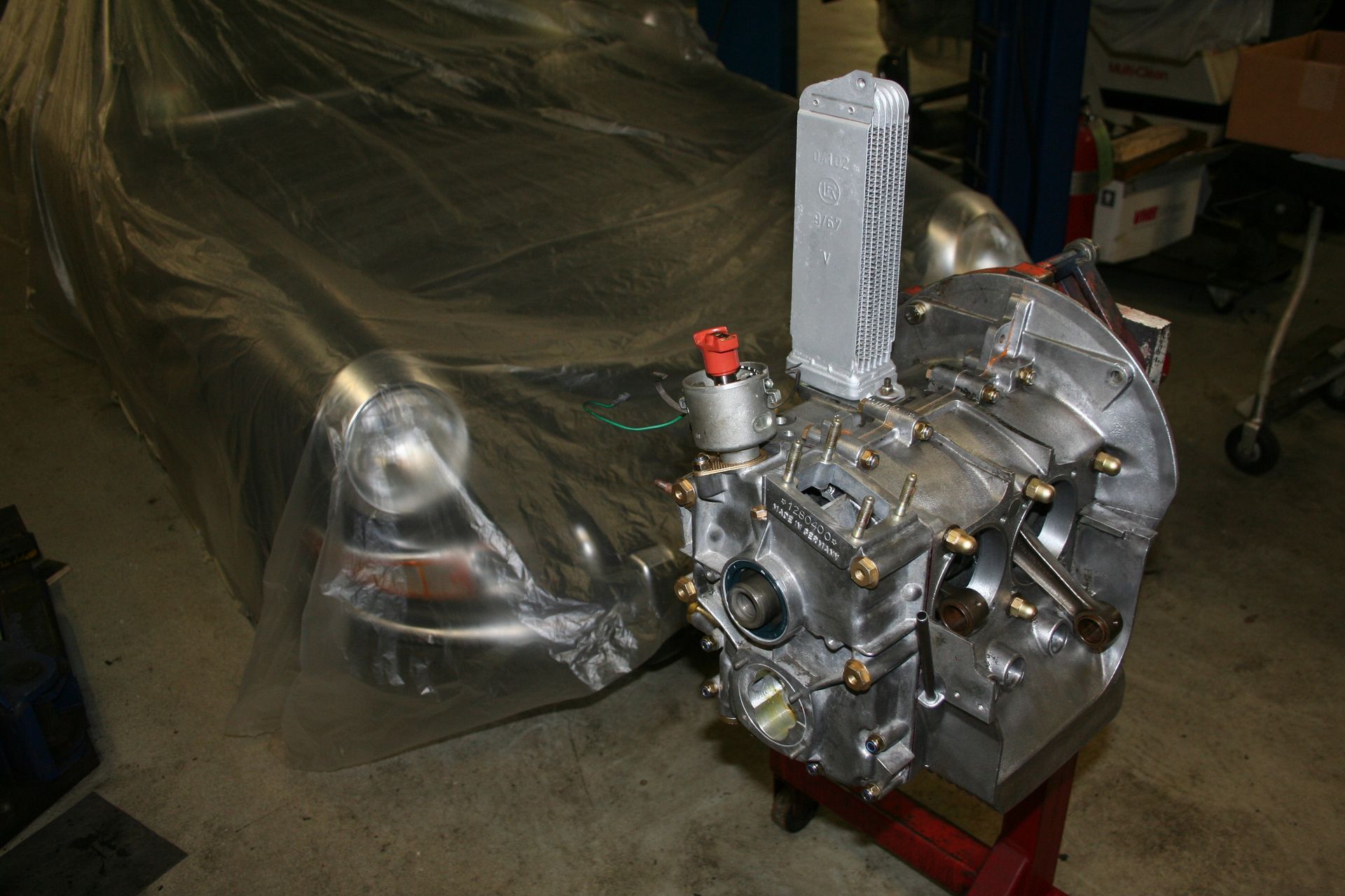 Silver engine block with attachments, likely for a vehicle, partially obscuring a car covered in plastic wrap. | Autobahn Performance Inc