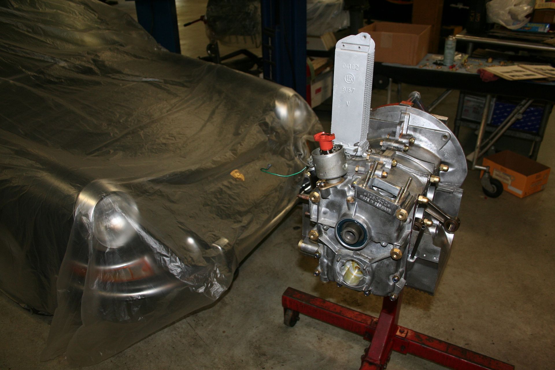 A silver engine on a red stand next to a car covered in plastic wrap in a workshop. | Autobahn Performance Inc