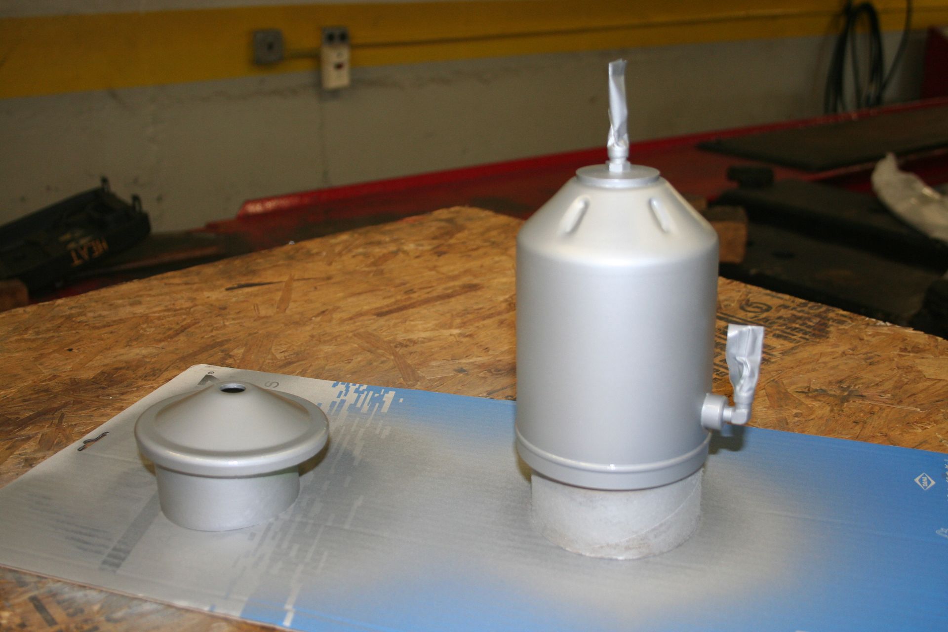 Two silver painted metal objects on a wooden surface. One is a cylinder, the other a cone-shaped lid. | Autobahn Performance Inc