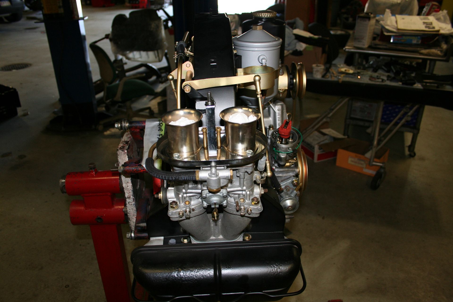 Engine with dual carburetors, gold-colored parts, on a red stand in a workshop. | Autobahn Performance Inc