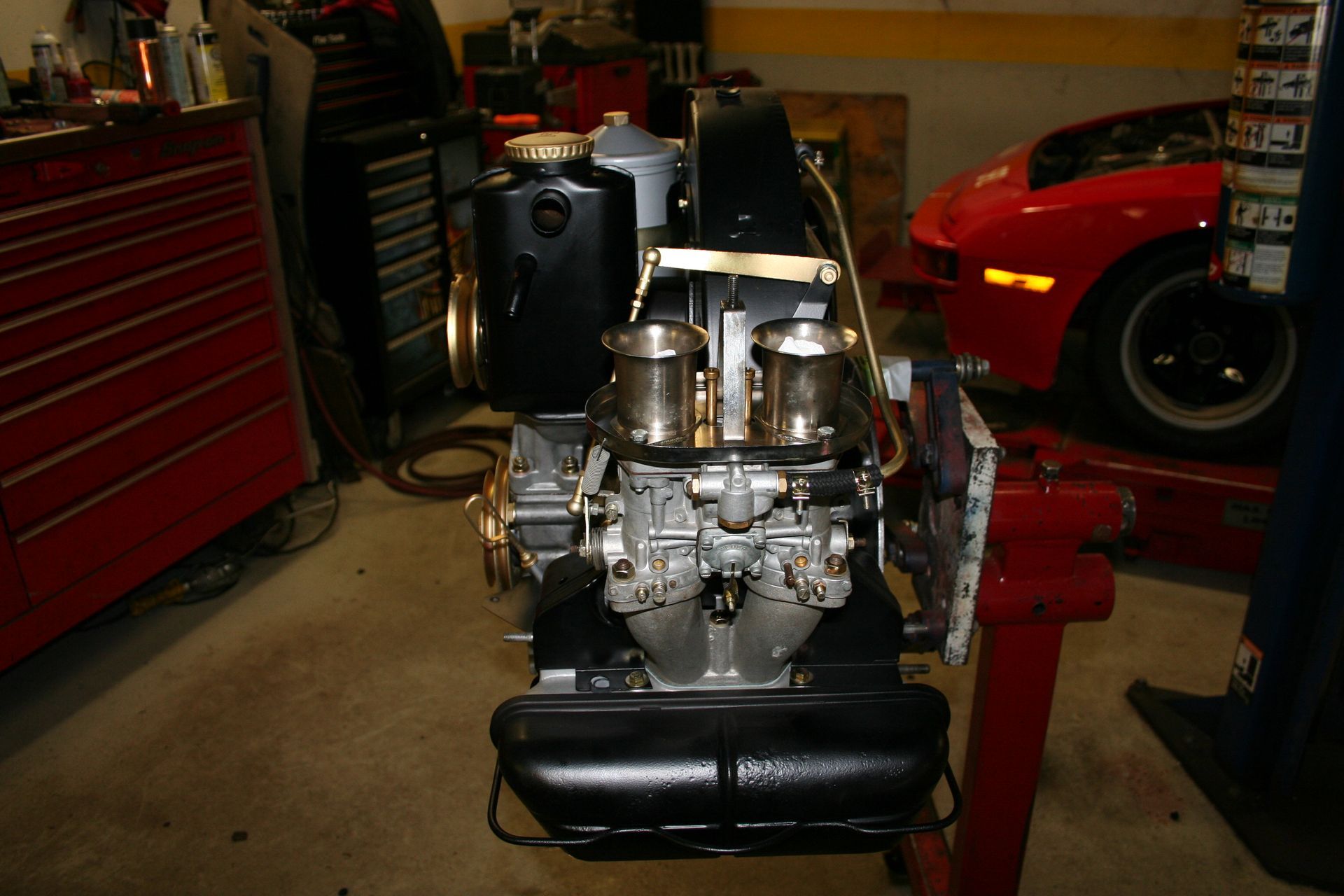 Black engine with silver carburetors, oil tank, and other components on a stand, red toolbox in background. | Autobahn Performance Inc