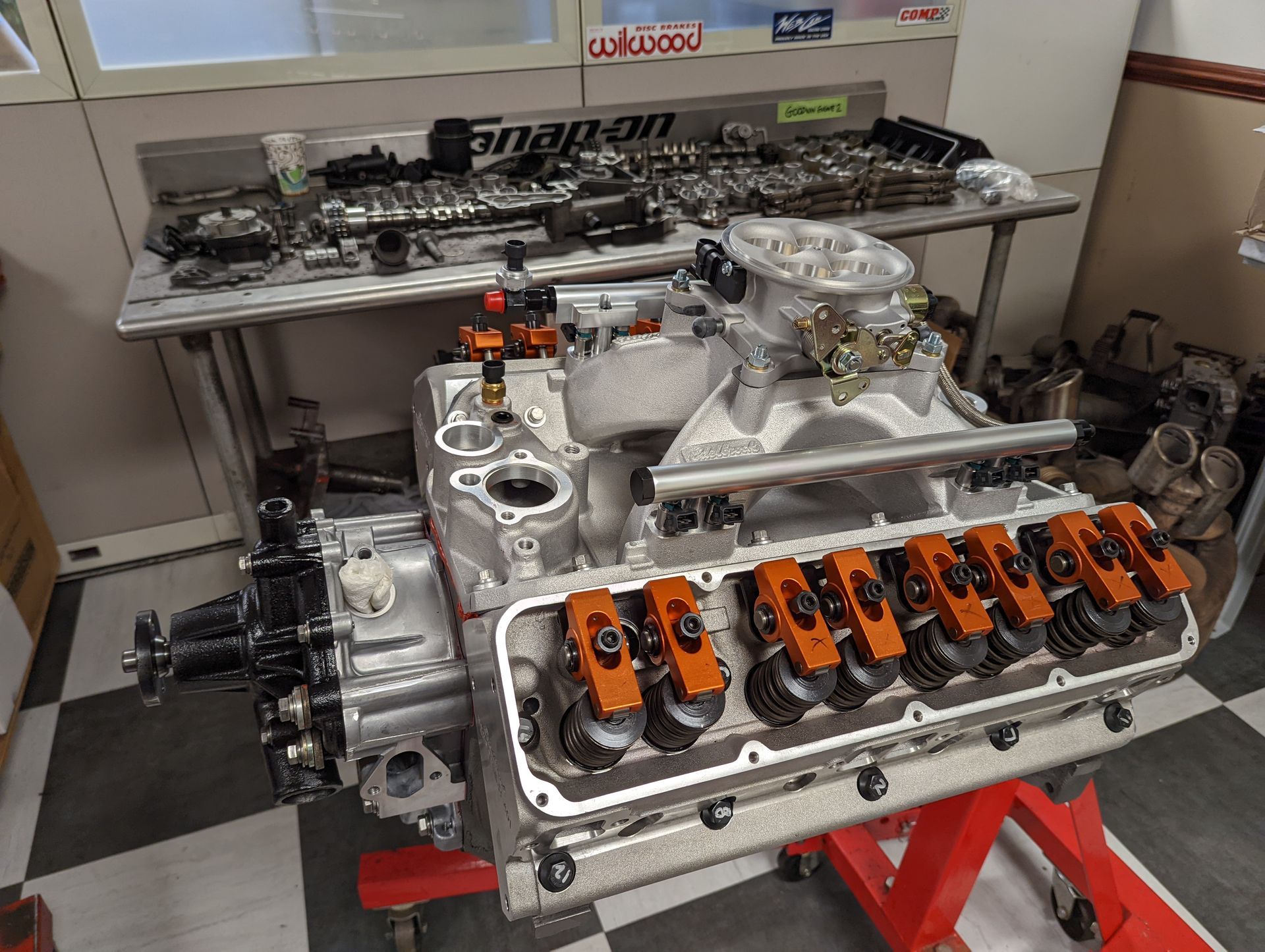 Assembled car engine with orange rocker arms, carburetor, and parts on a nearby workbench. | Autobahn Performance