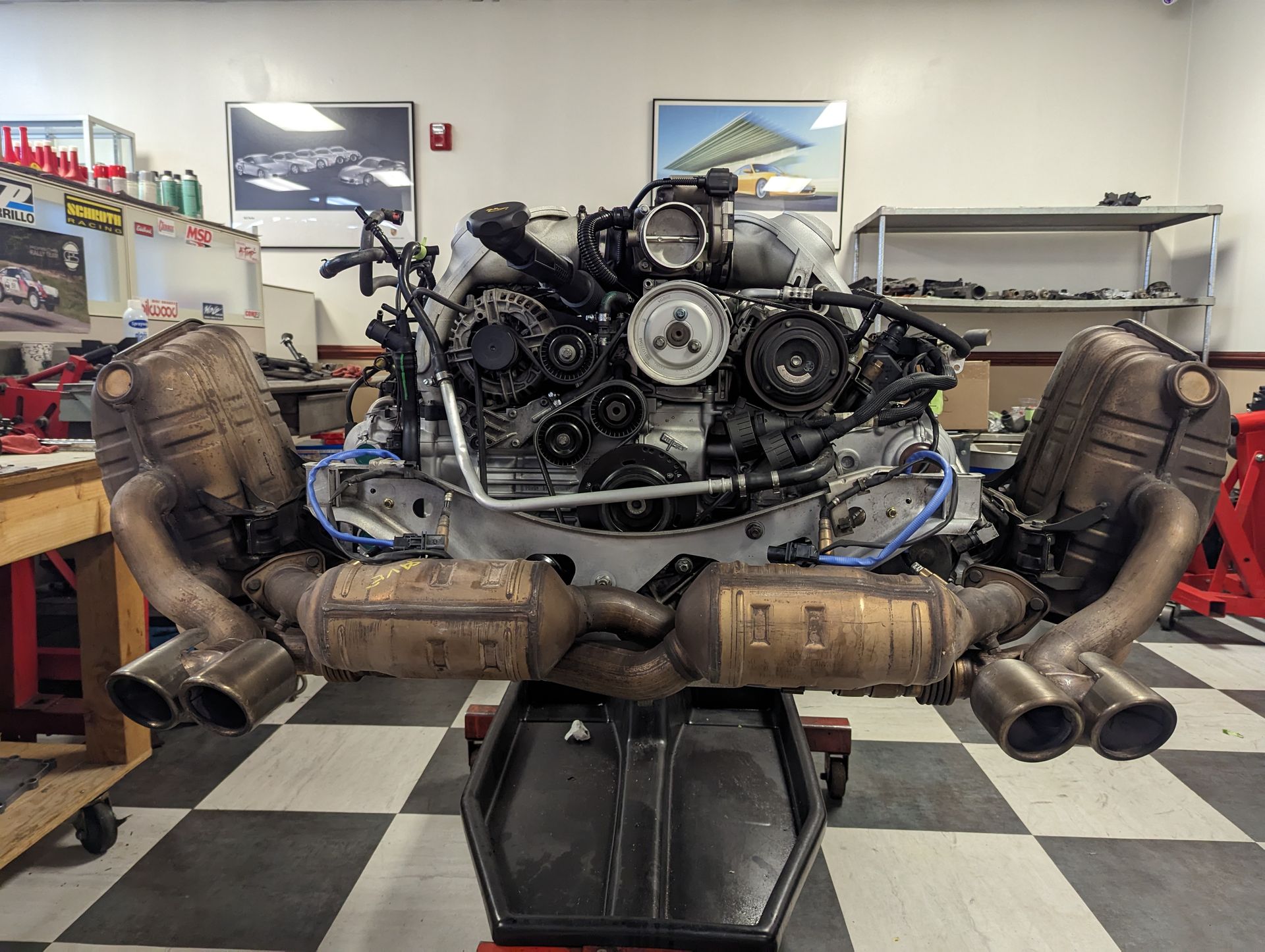 Porsche engine with exhaust system, sitting on a stand in a workshop. Gray, black, and silver components. | Autobahn Performance