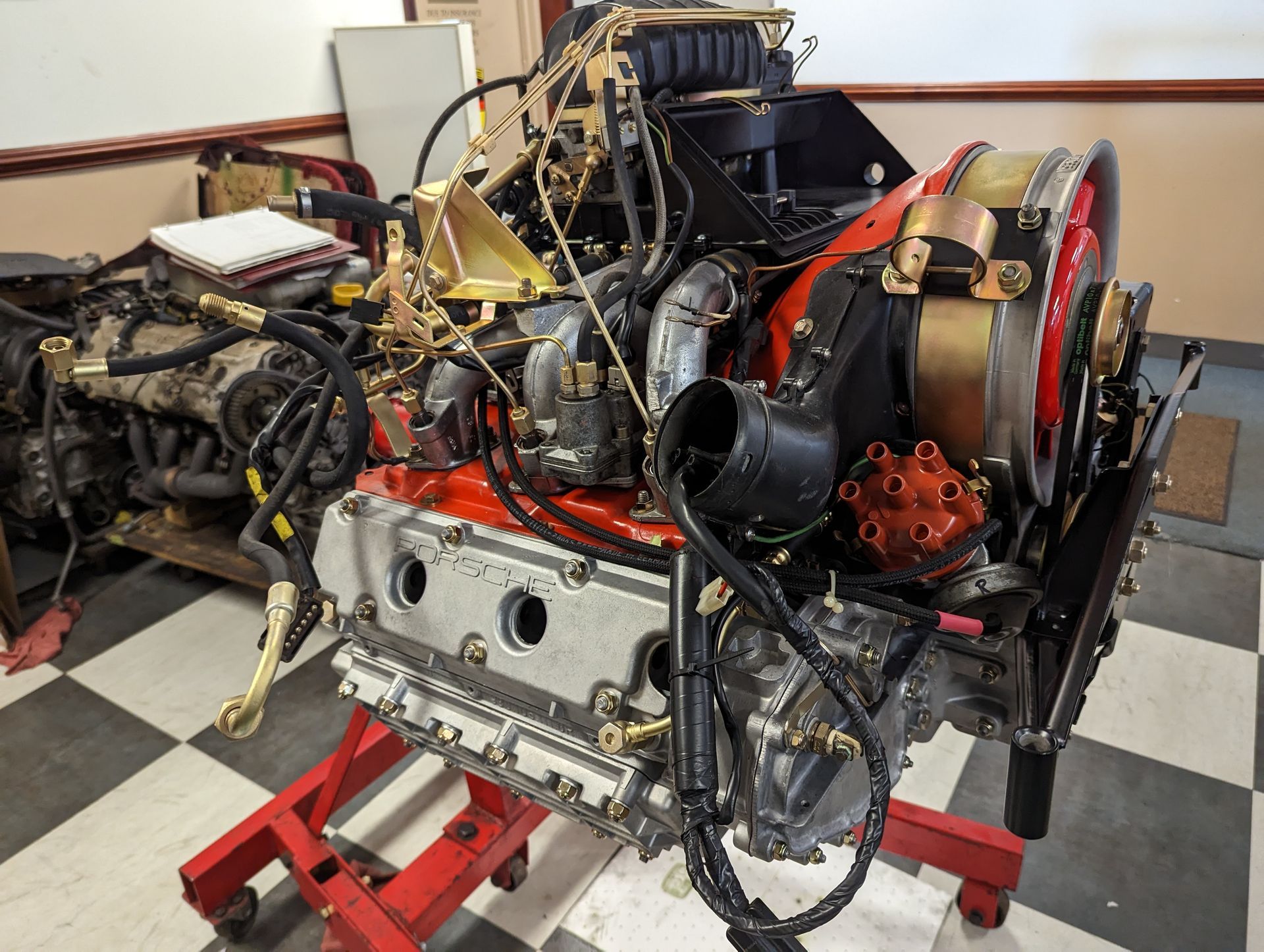 Porsche engine on a red stand, black hoses and wires, gold and red accents, in a workshop. | Autobahn Performance Inc