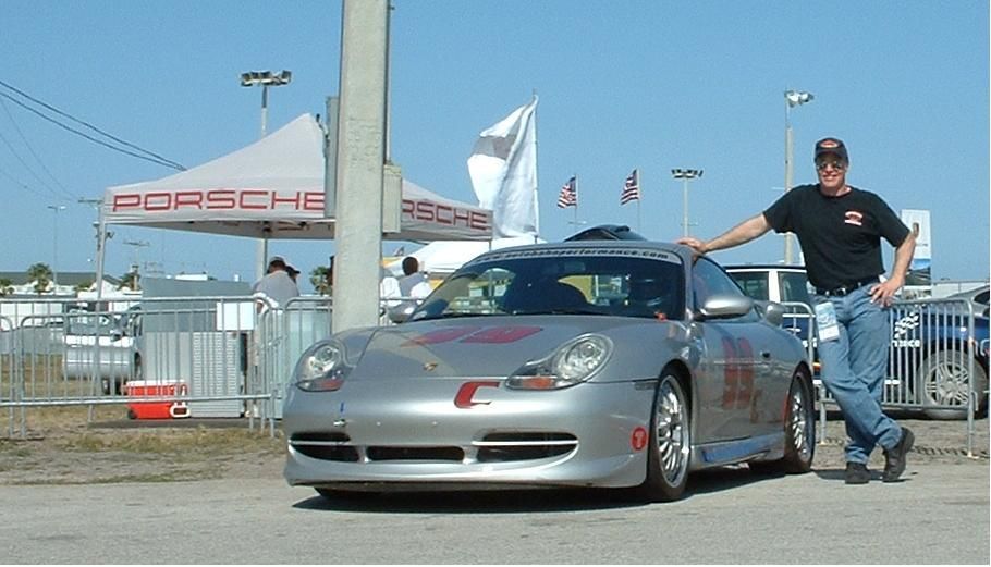 Man poses next to a silver Porsche sports car in front of a Porsche tent at an outdoor event. | Autobahn Performance Inc