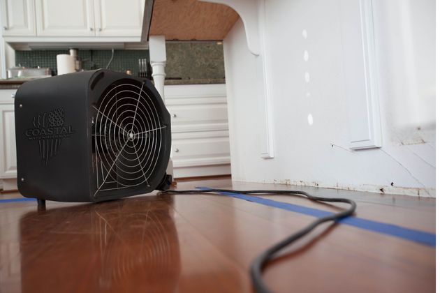 A black fan is sitting on a wooden floor in a kitchen.